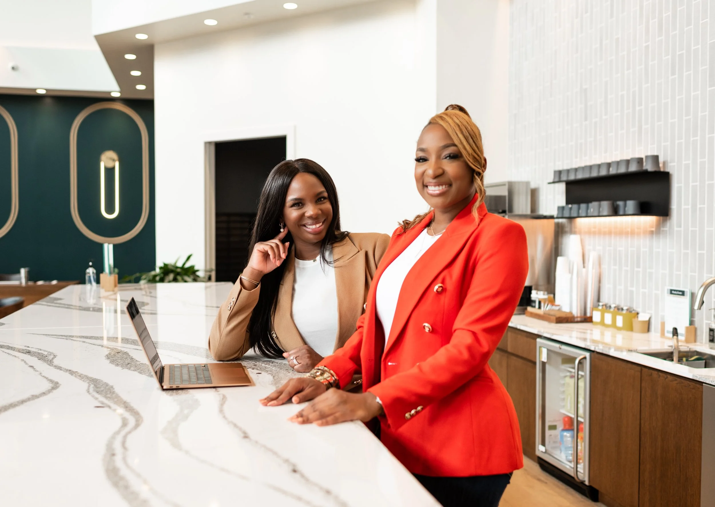 Two women smiling at the counter in a modern cafe or office kitchen, one wearing a beige blazer and the other in a red blazer, with a laptop on the counter.