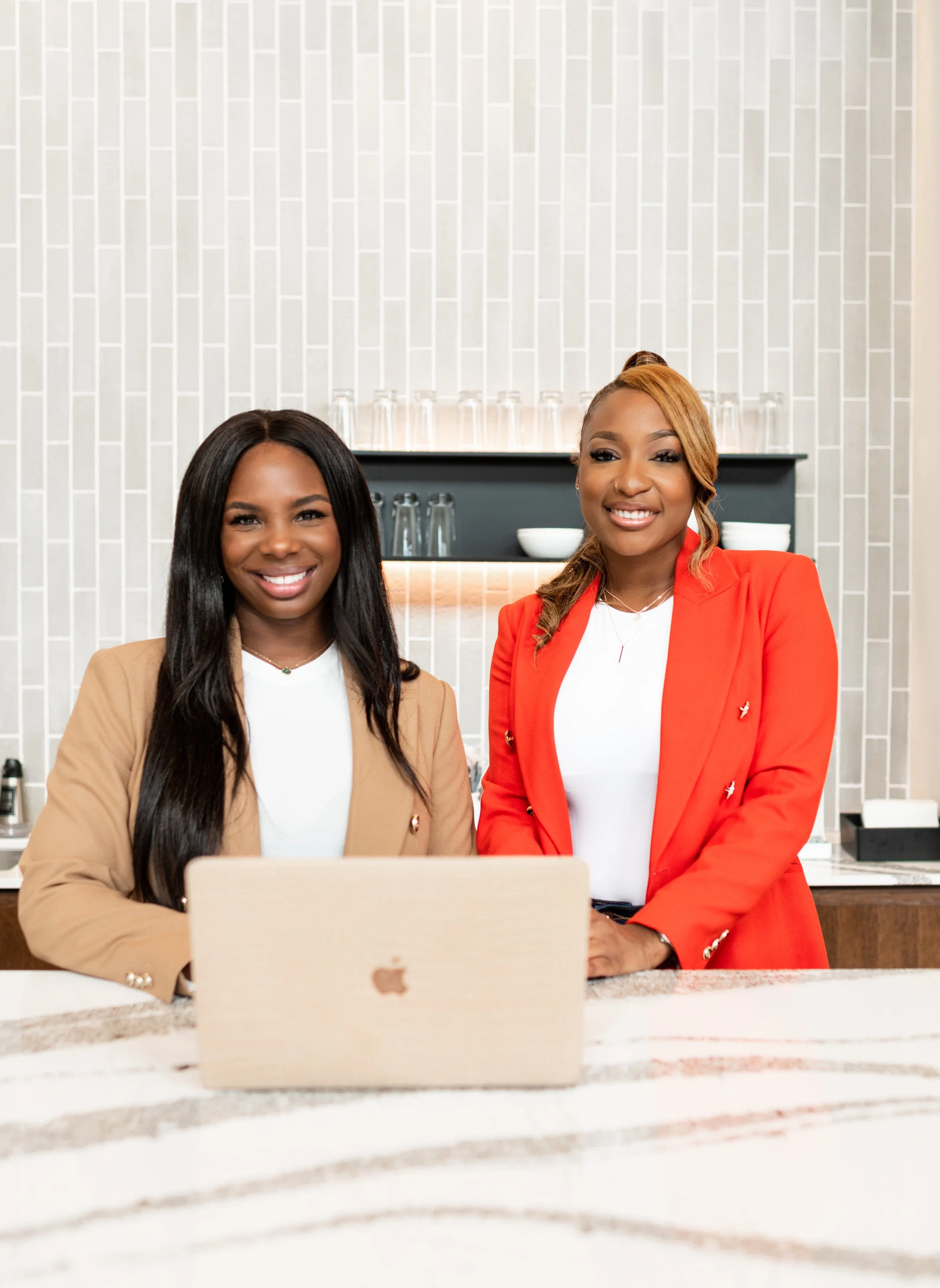 Two women standing behind a counter with a laptop, smiling, in a modern kitchen or cafe setting.