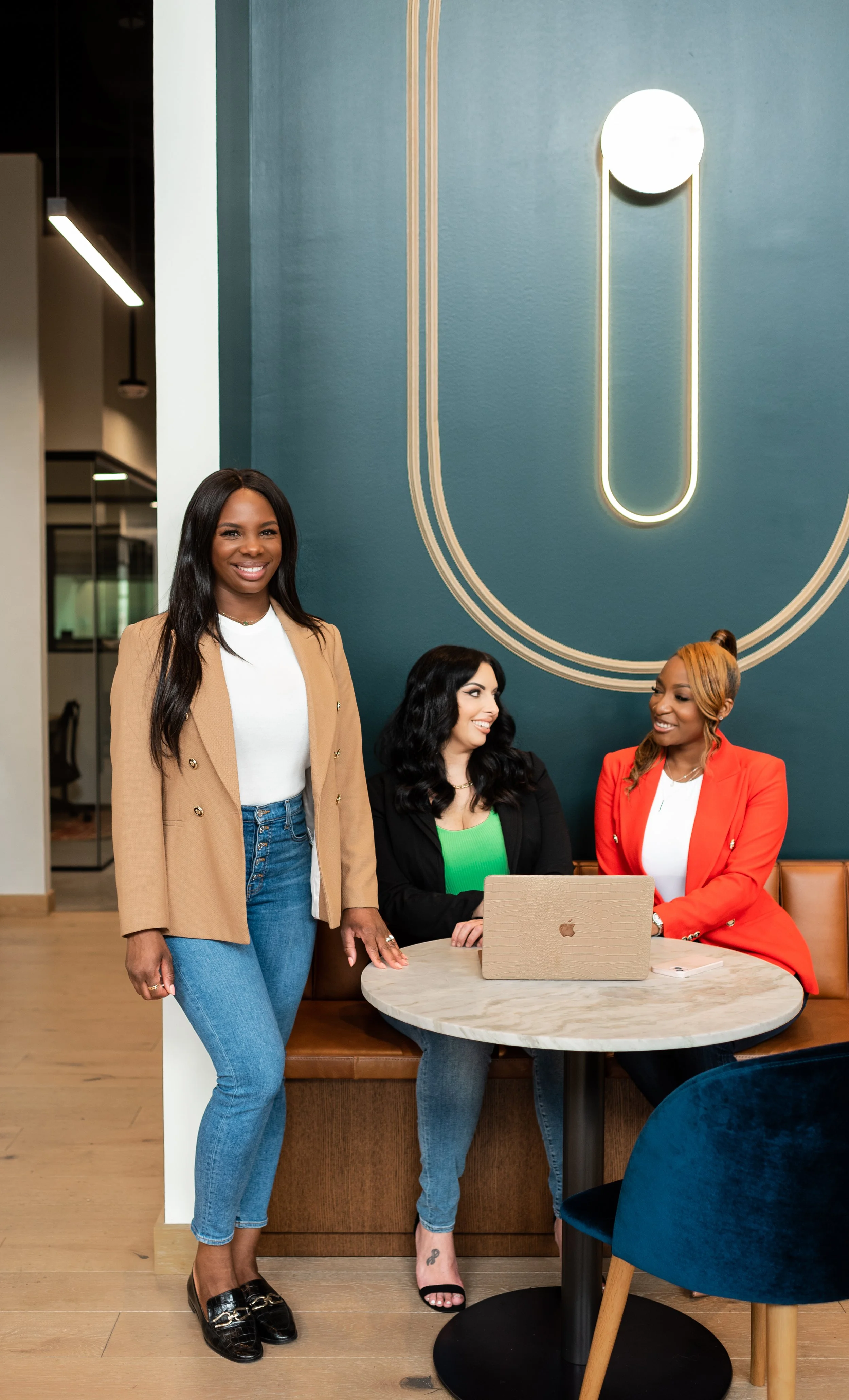 Three women in a modern office lounge area, one standing and two seated at a round marble table with a laptop, smiling and engaging in conversation.