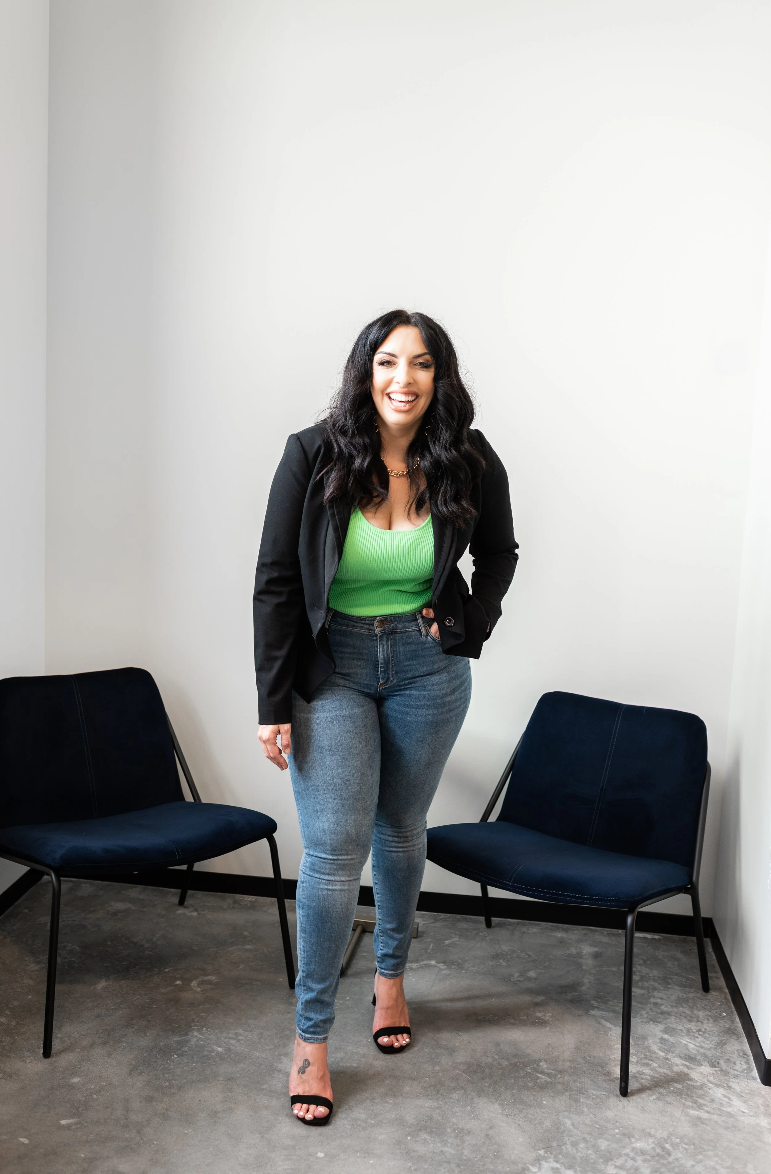 A woman standing in a room with white walls and concrete floor, smiling and posing for the camera, flanked by two black chairs.