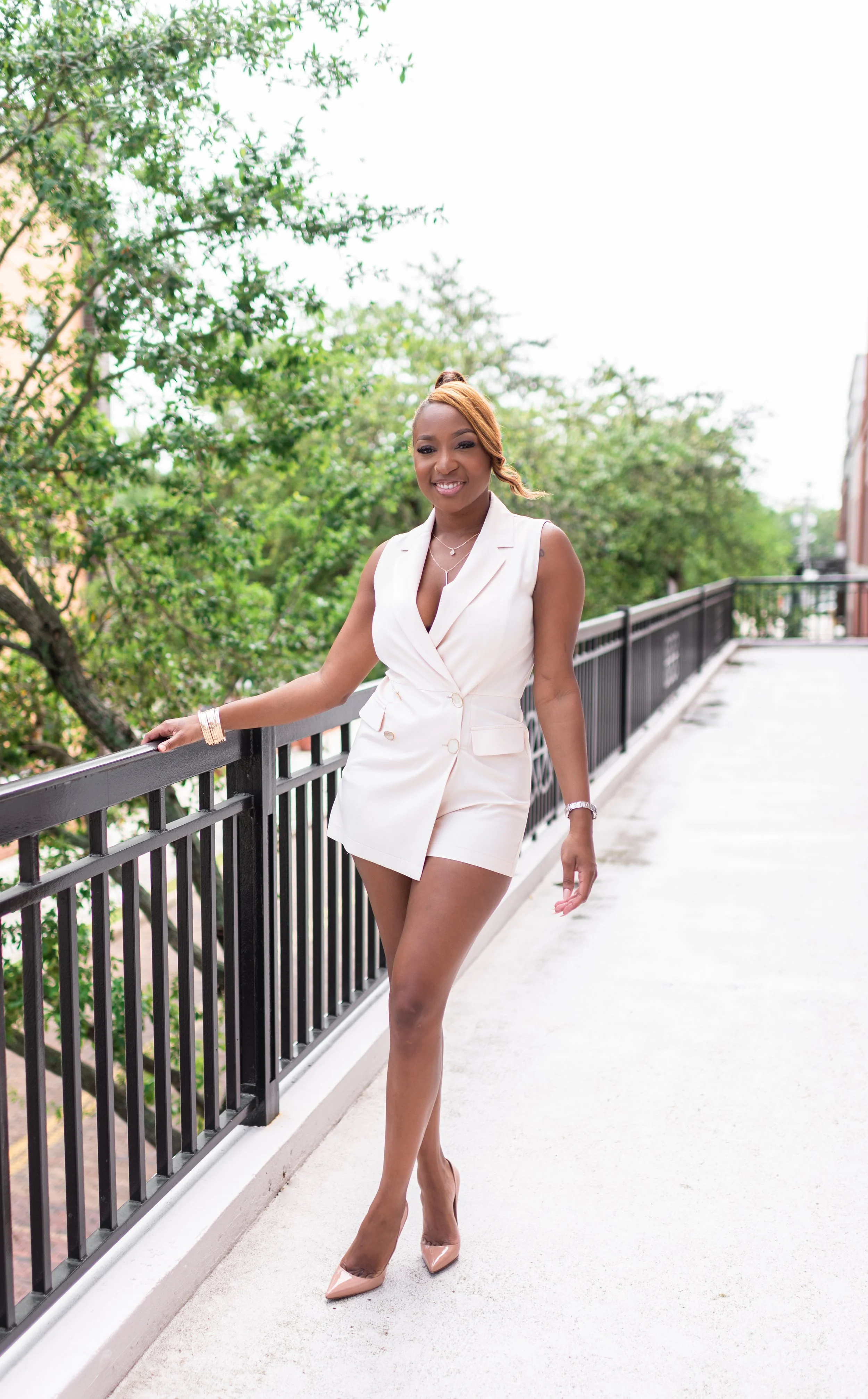 A woman in a white sleeveless blazer and shorts outfit, wearing beige heels, standing on a balcony with a black railing, smiling at the camera with green trees in the background.