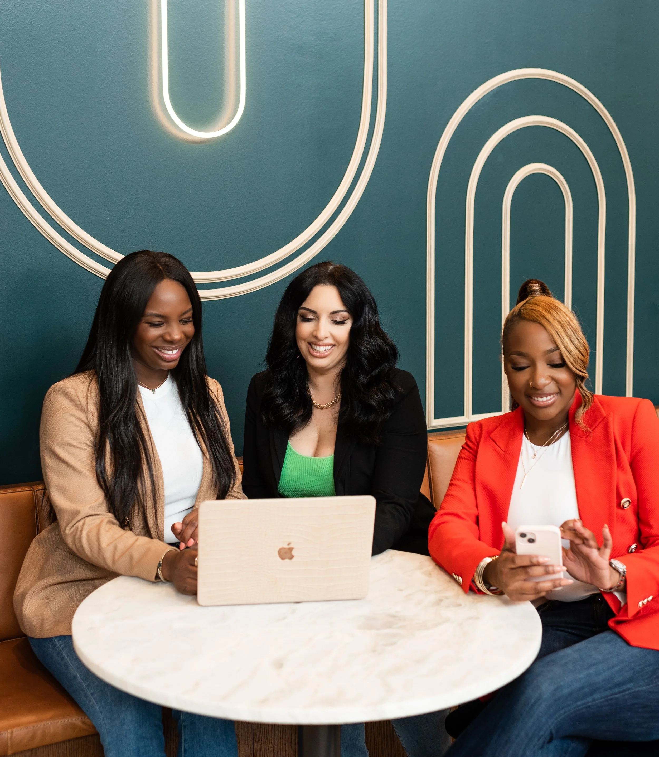 Three women sitting at a marble table in a cafe, looking at a laptop and a smartphone, smiling, against a teal wall with modern white light art.