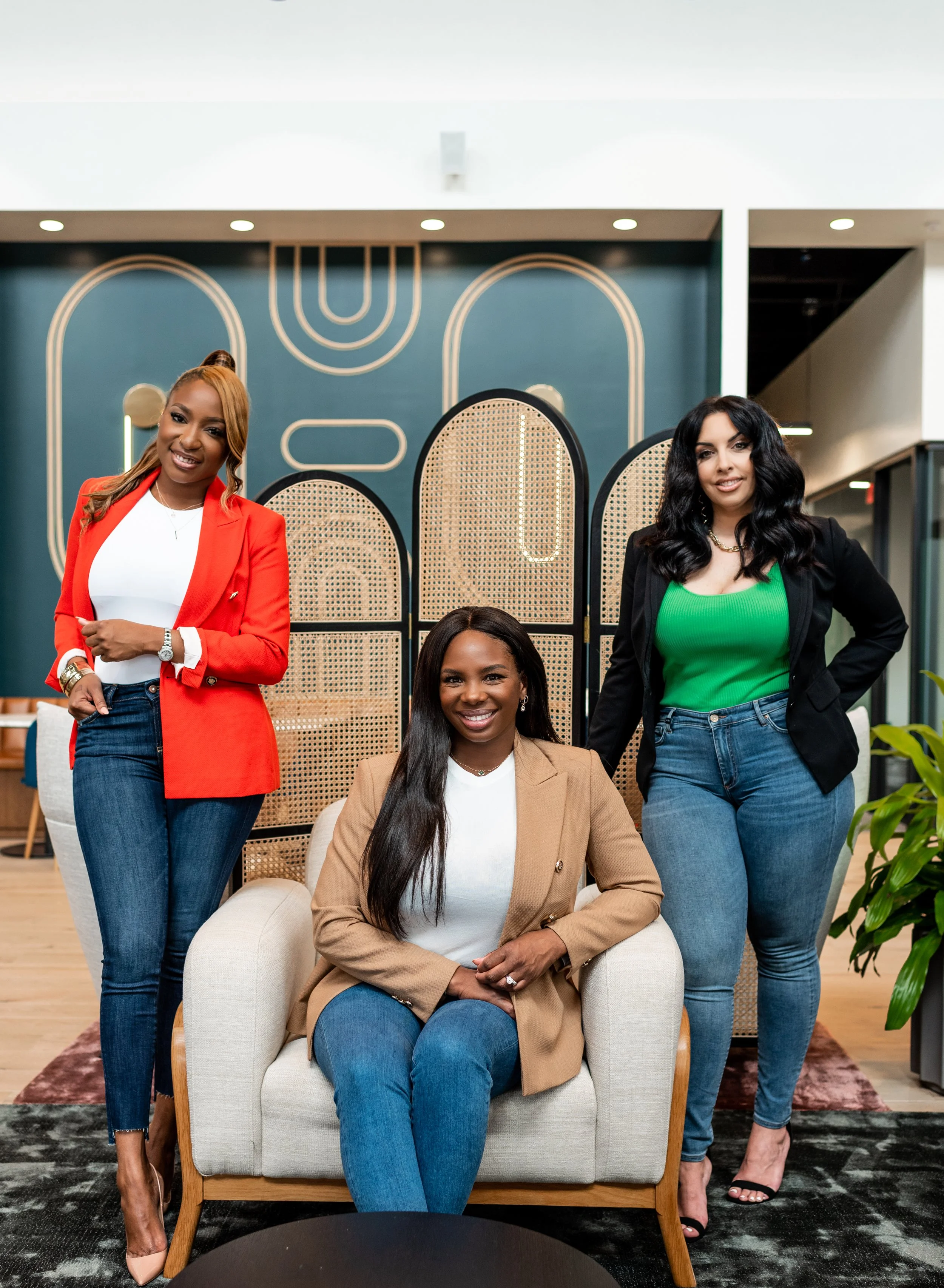 Three women in business casual attire posing in a modern office space with decorative screens and art on the wall.