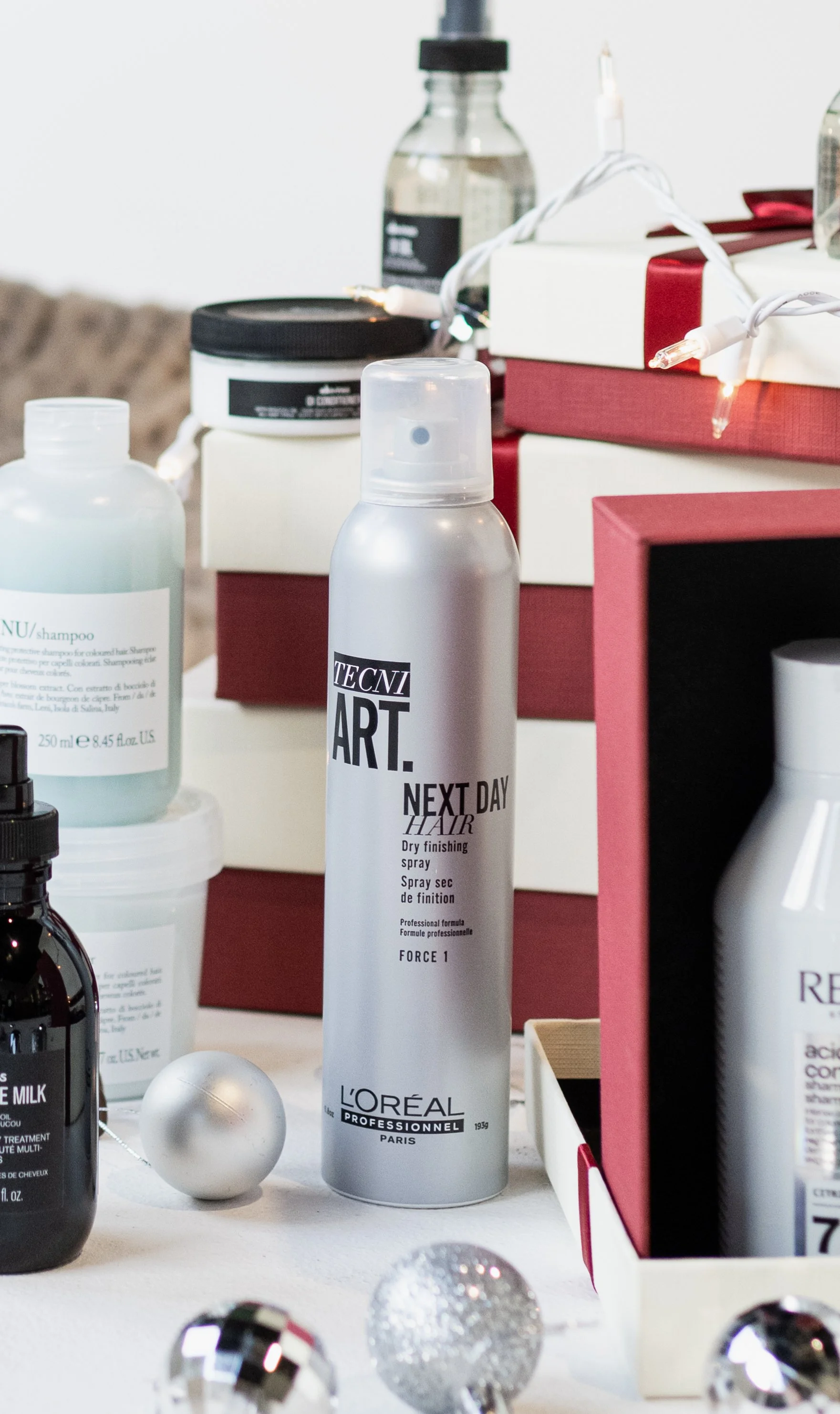 A silver can of L'Oréal Professionnel Next Day Hair dry finishing spray among various beauty products, Christmas ornaments, and gift boxes on a white surface.