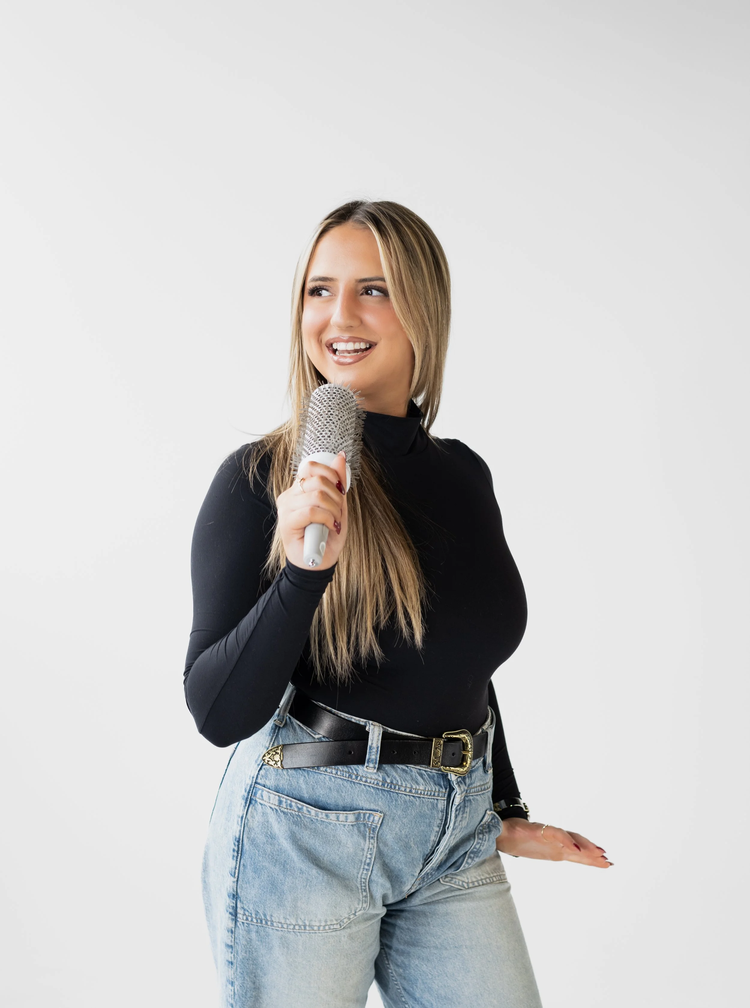 A young woman with long blonde hair, wearing a black long-sleeve top and light blue jeans, holding a microphone and smiling, against a plain white background.