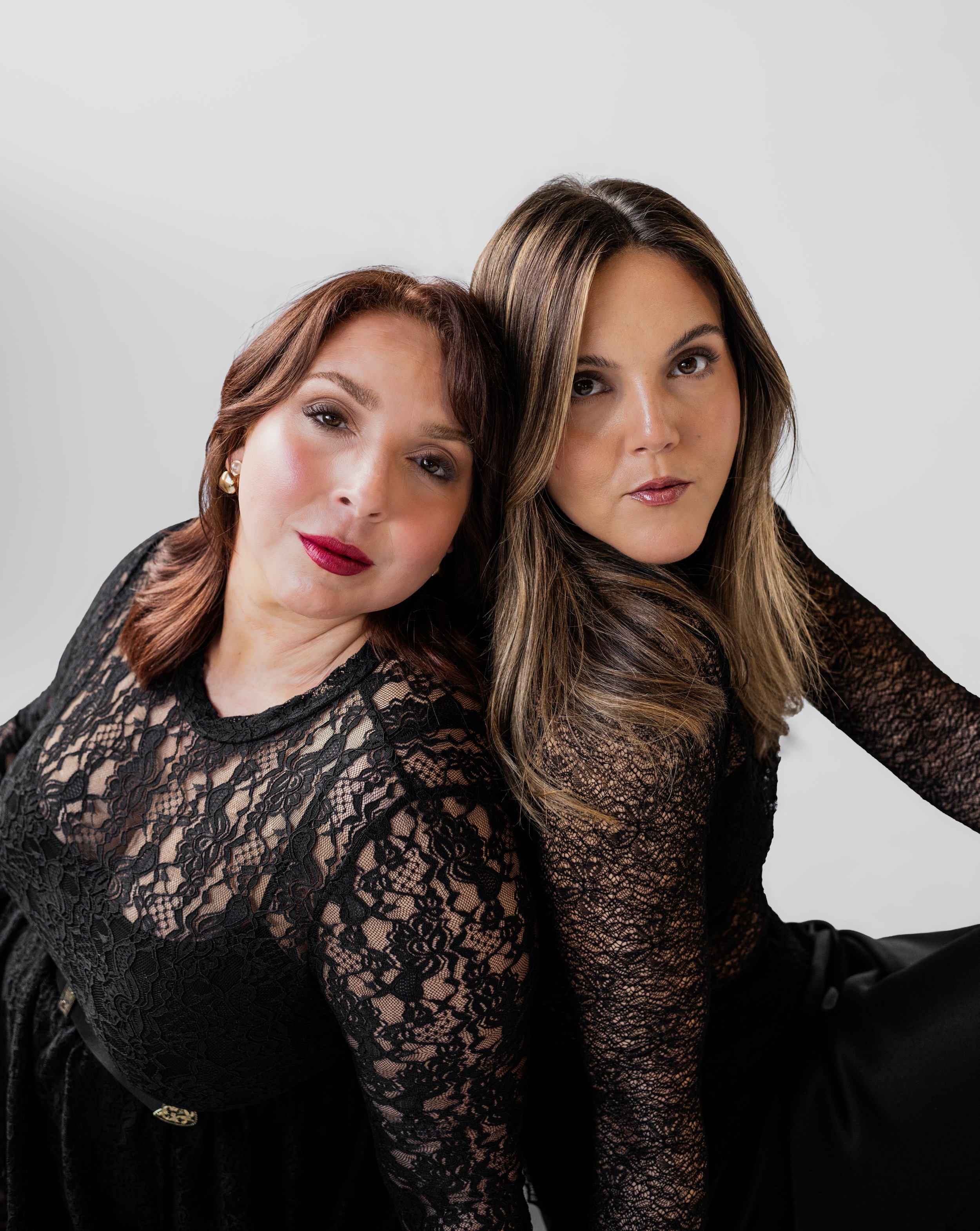 Two women in black lace dresses sitting close together, facing the camera.