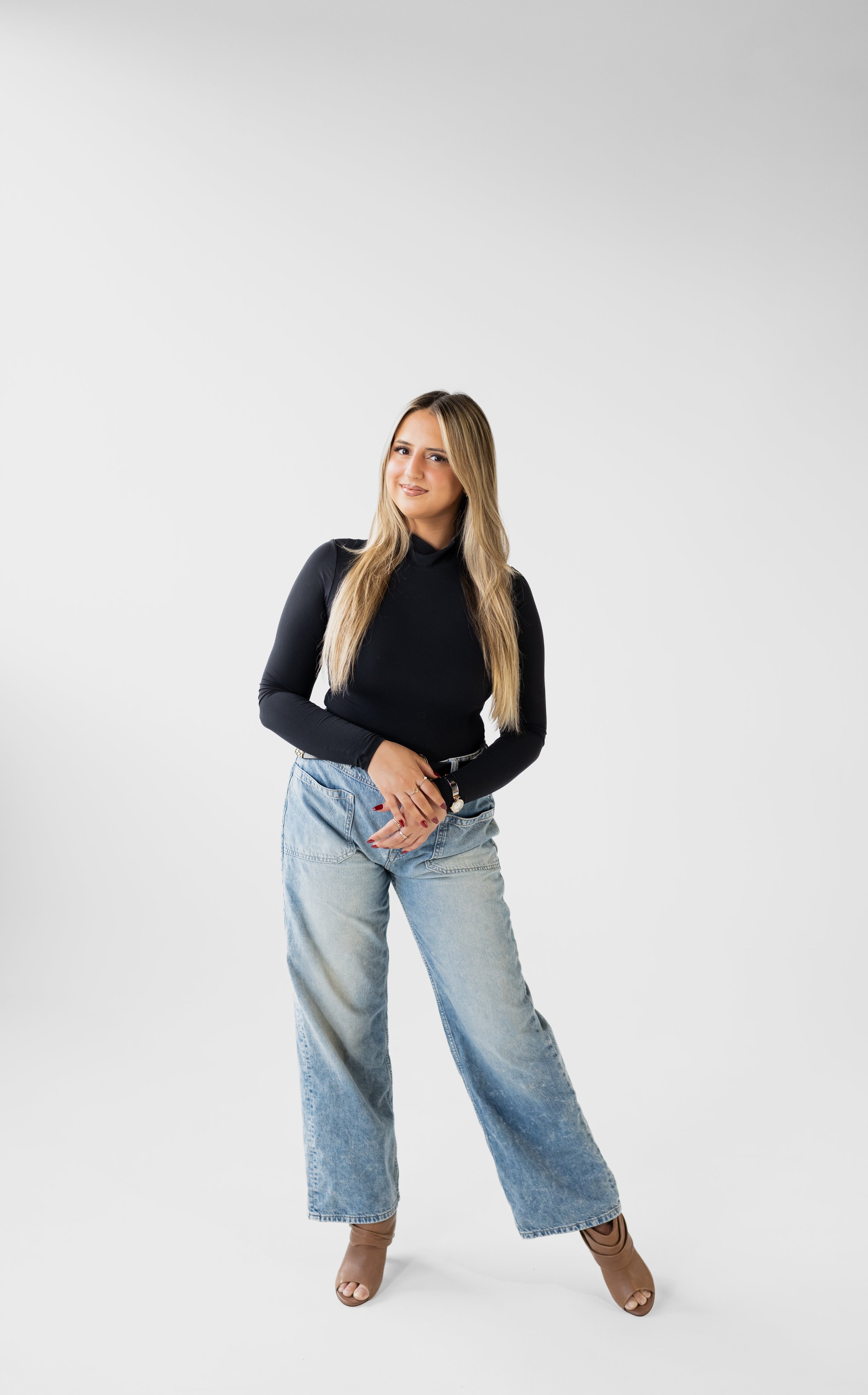 A woman with long blonde hair, wearing a black turtleneck and light blue wide-leg jeans, standing against a plain white background with a slight smile and confident pose.