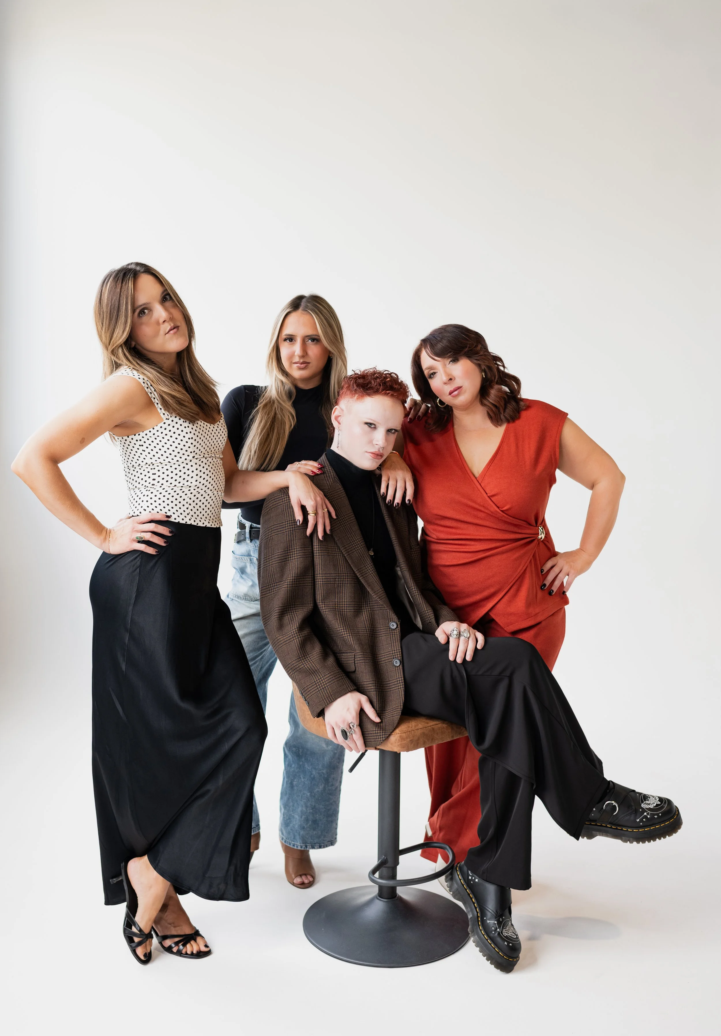 Four stylish women posing together on a white background, with one seated on a stool and the other three standing around her.