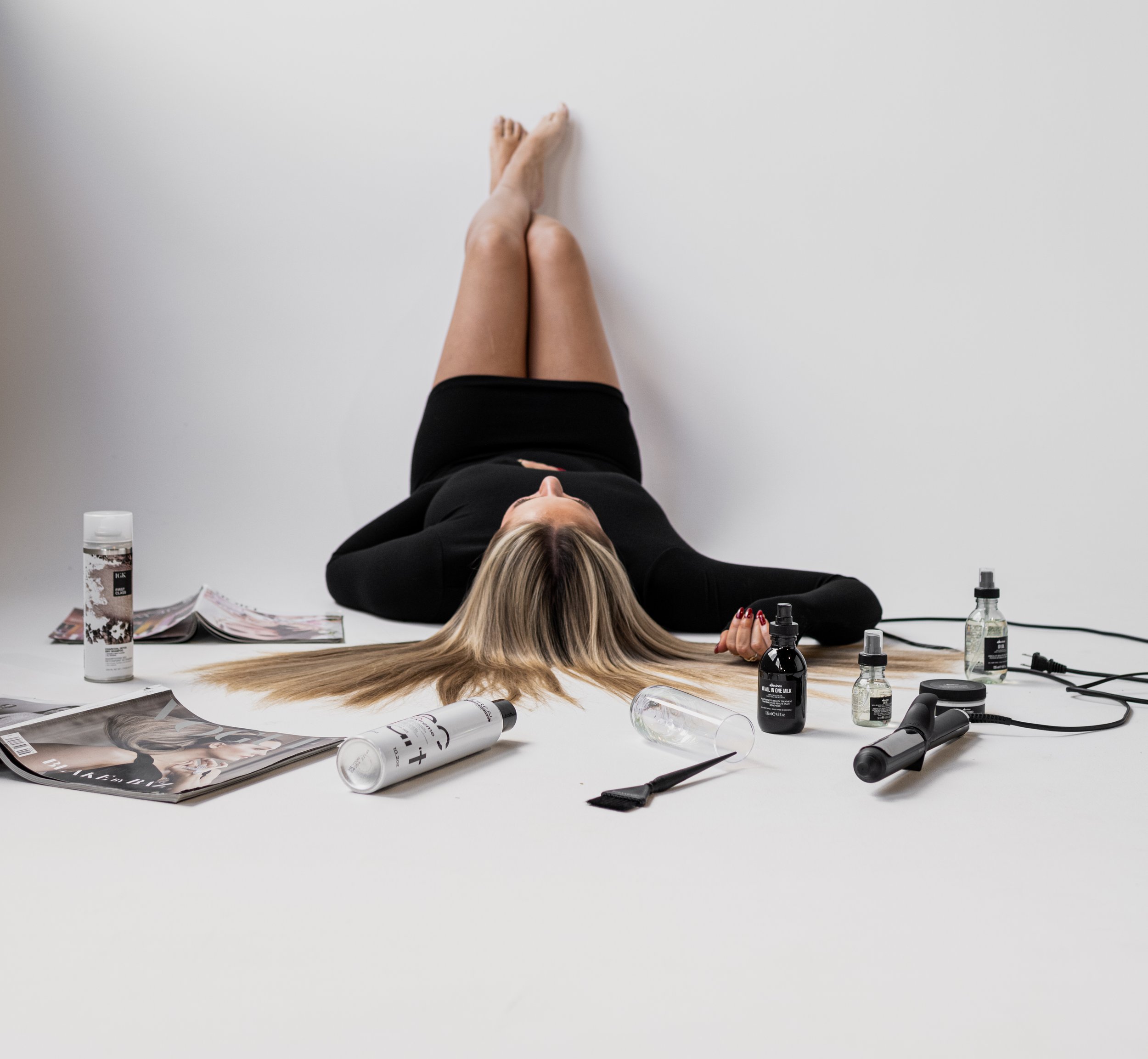 A woman lying on the floor surrounded by hair styling products and magazines, with her long blonde hair spread out around her head.