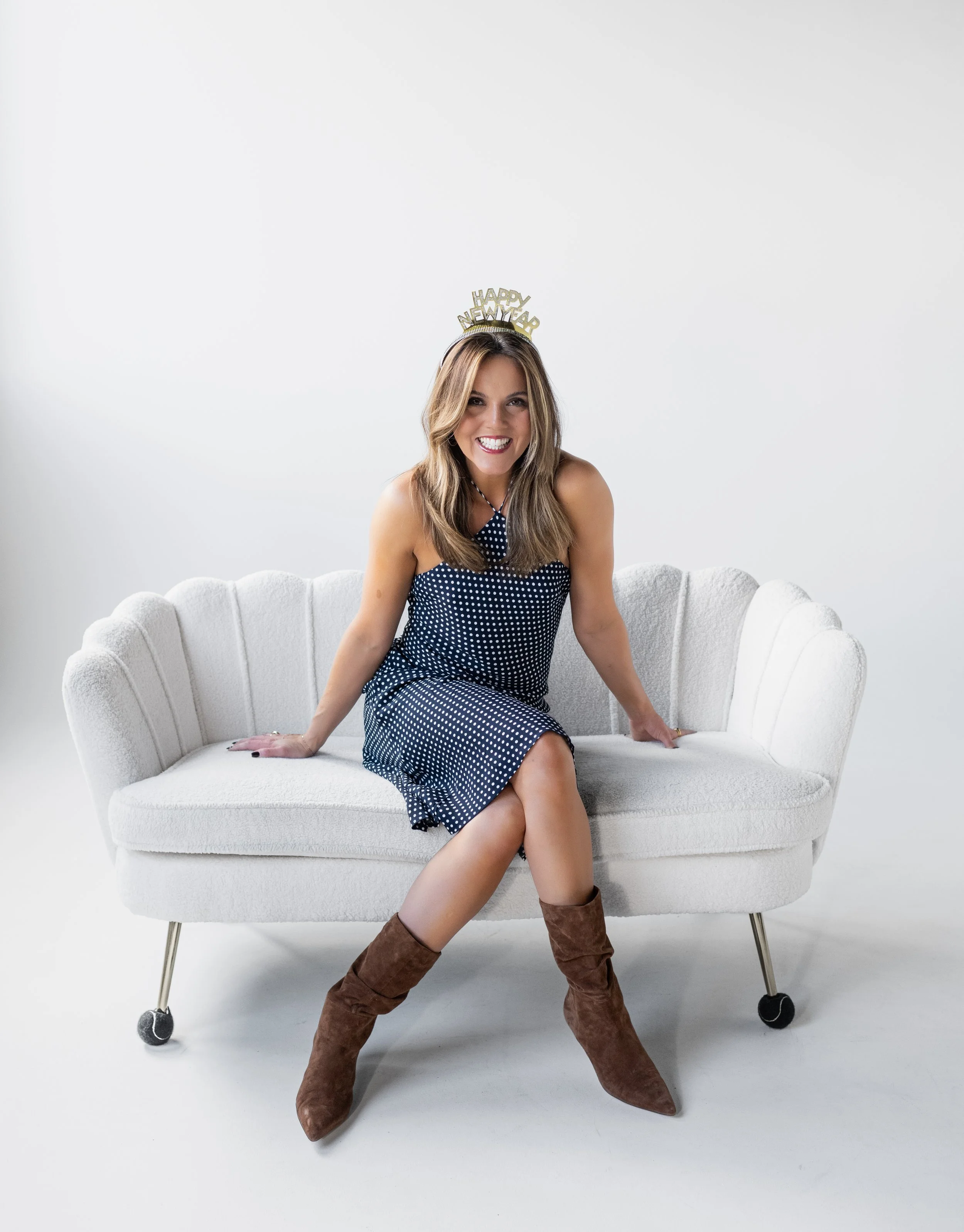 A woman in a polka dot dress and brown boots sitting on a white vintage sofa, wearing a "Happy New Year" headband, smiling at the camera against a white background.