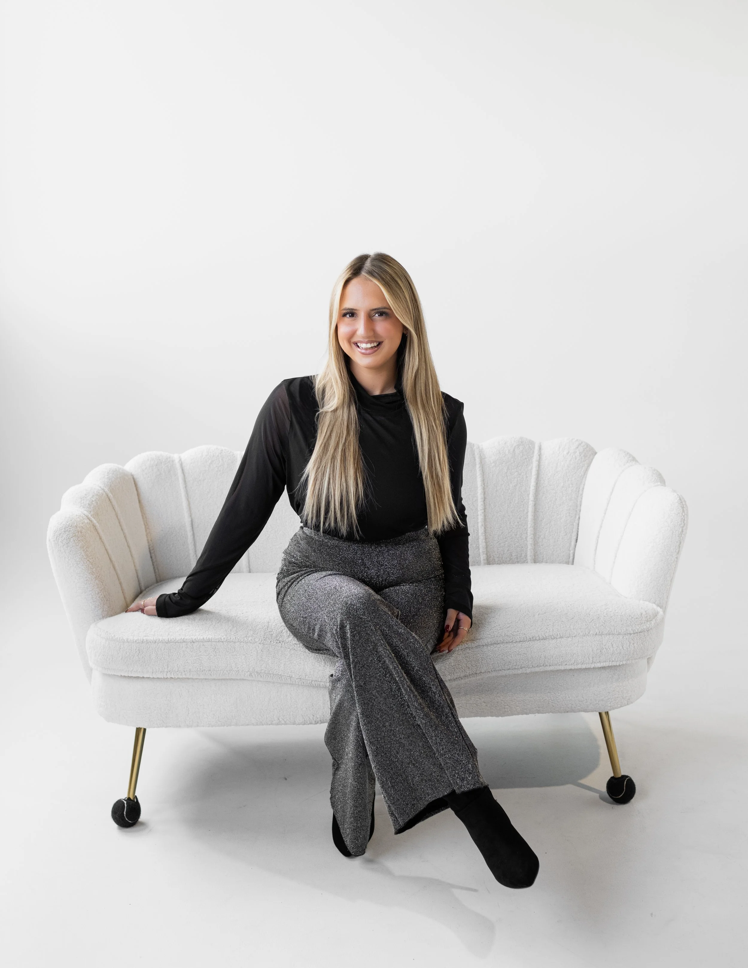 A woman sitting on a white, vintage-style sofa with gold legs and black casters, dressed in a black top and glittery gray pants, smiling against a plain white background.