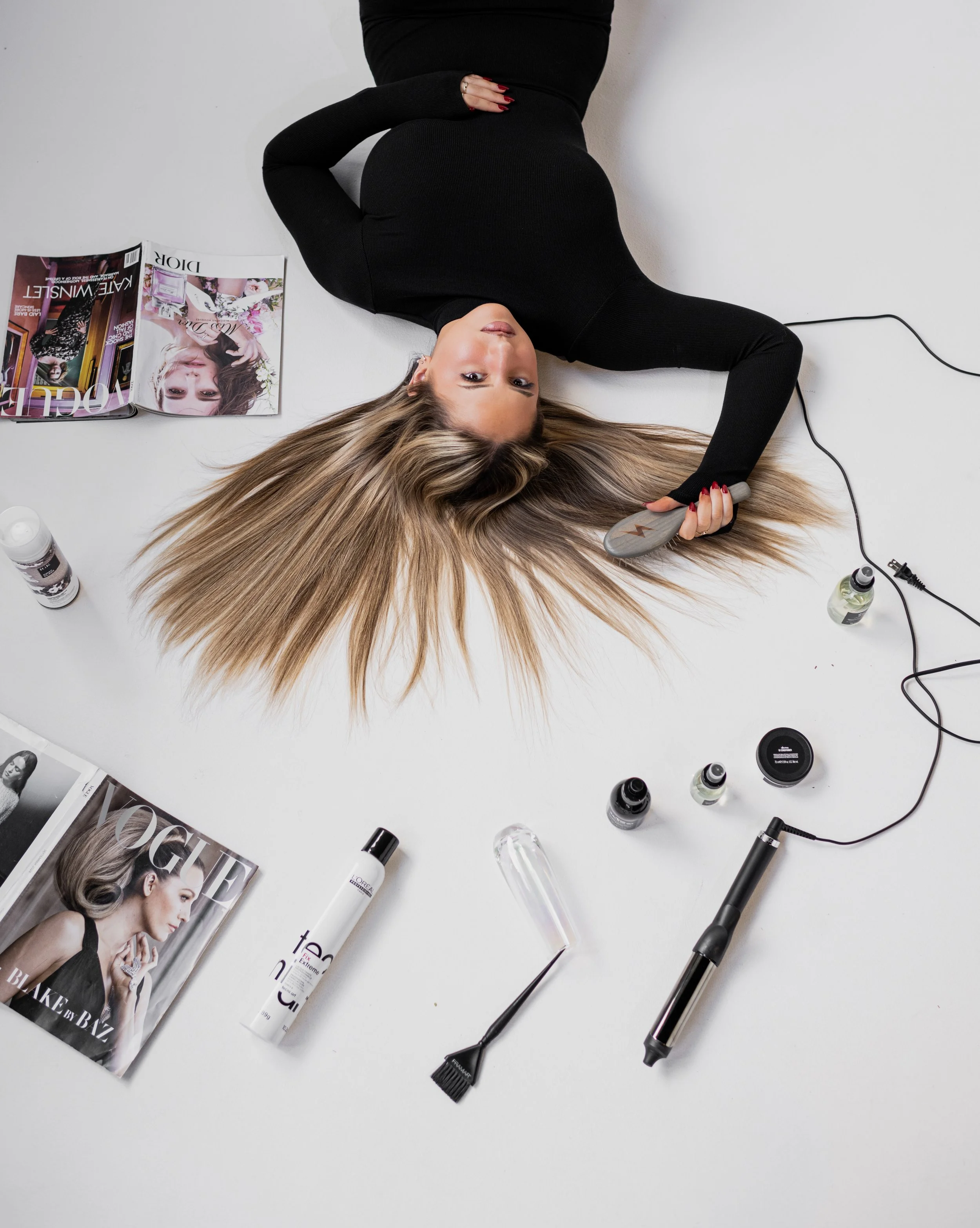 A woman lying on her back on a white surface, holding a hairbrush, surrounded by beauty and hair styling products and magazines.