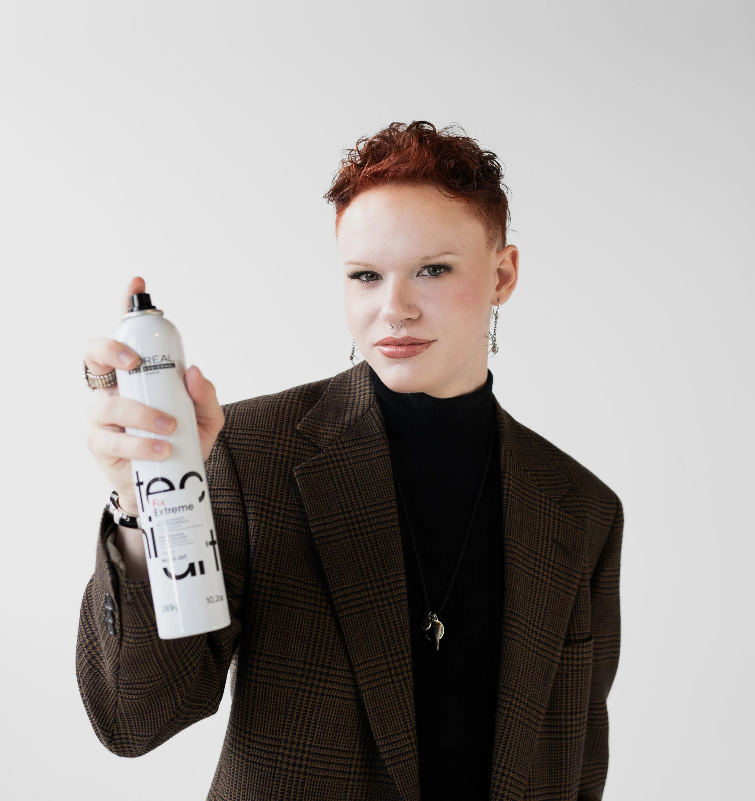 A woman with short red hair wearing a checkered blazer and black turtleneck holding a can of hairspray and looking at the camera against a plain white background.