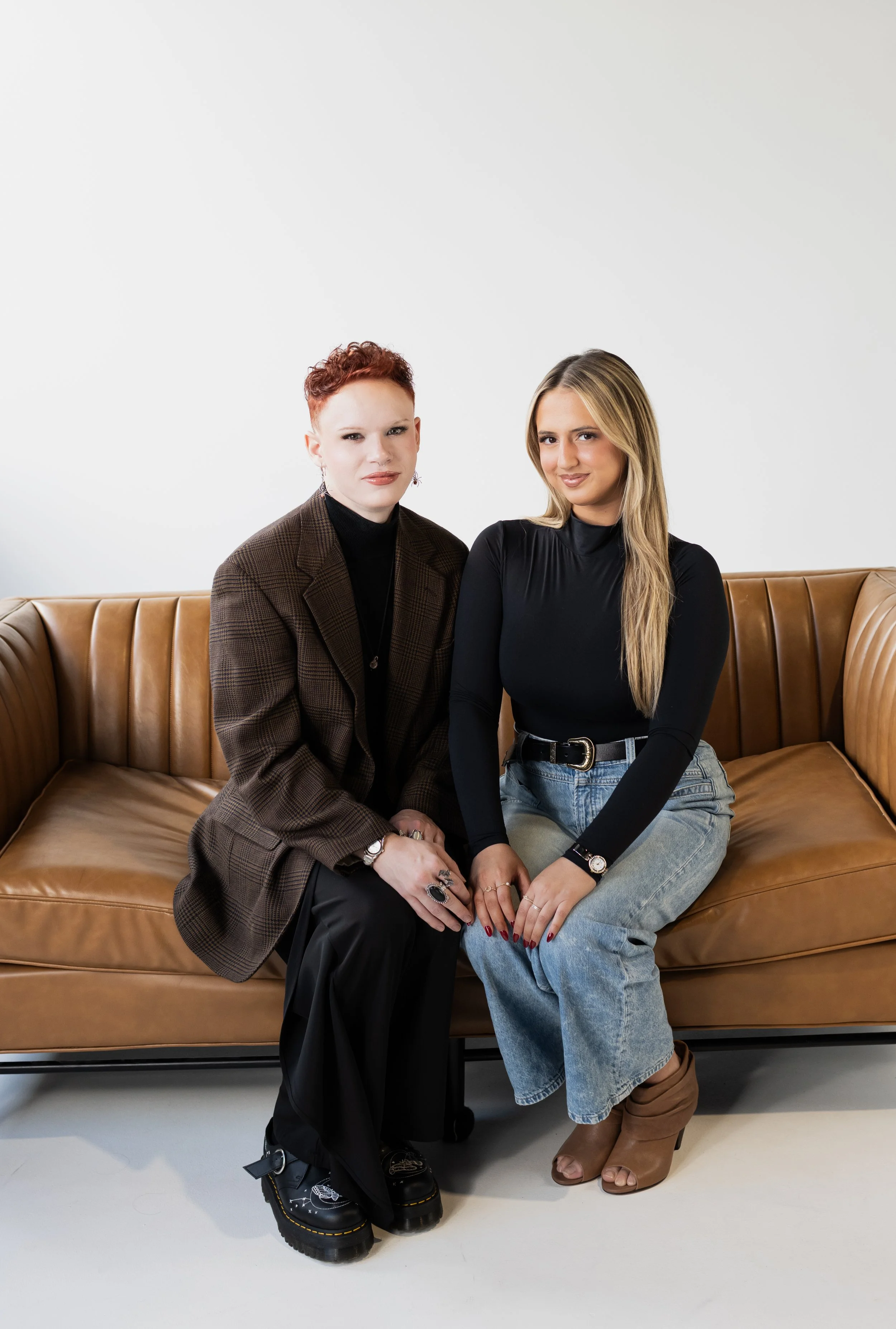 Two women sitting on a brown leather couch against a white background, both wearing black tops and casual pants, with watches and rings, posing for the photo.