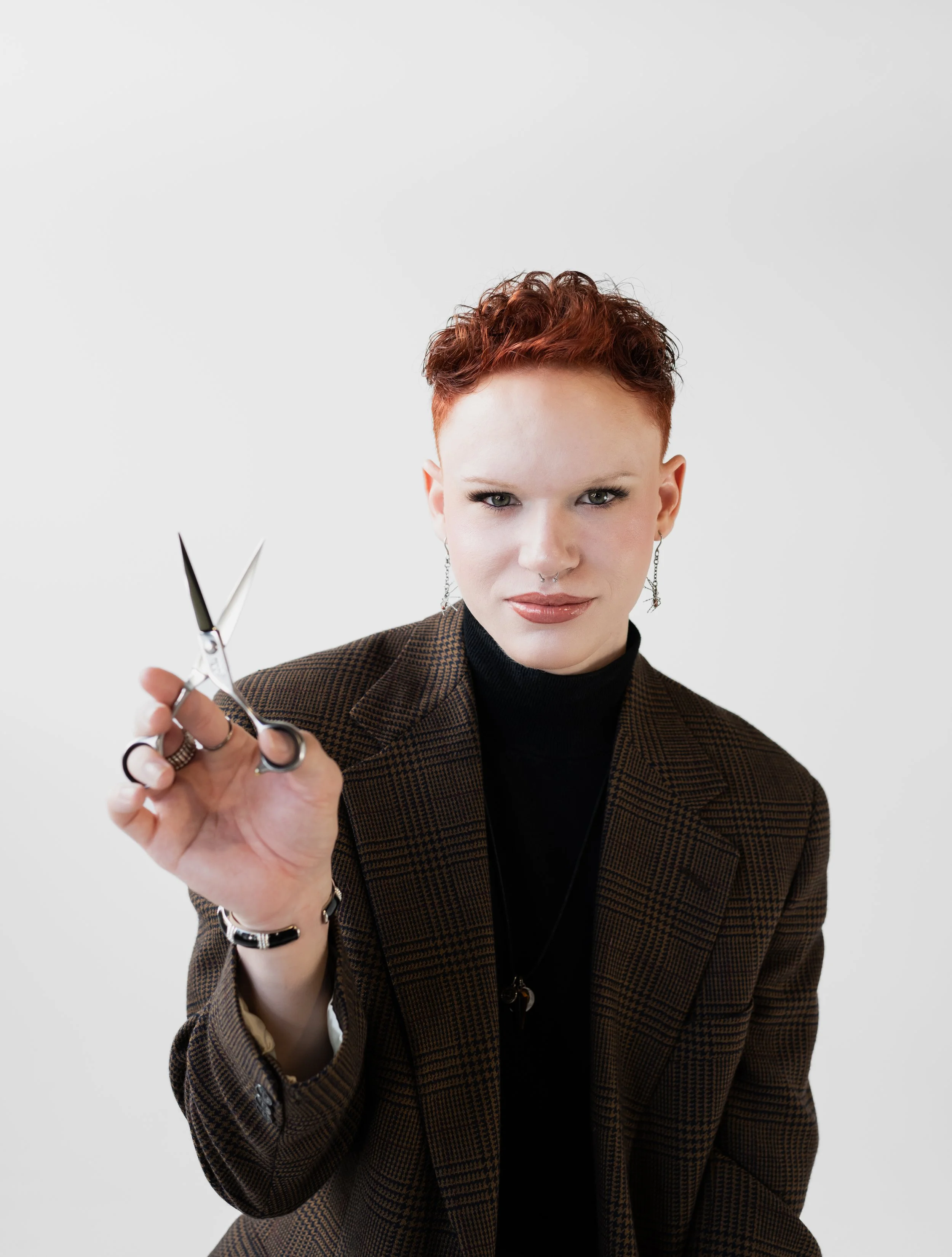 A woman with short red hair holding a pair of scissors toward the camera, wearing a brown checkered blazer and black turtleneck, against a plain white background.
