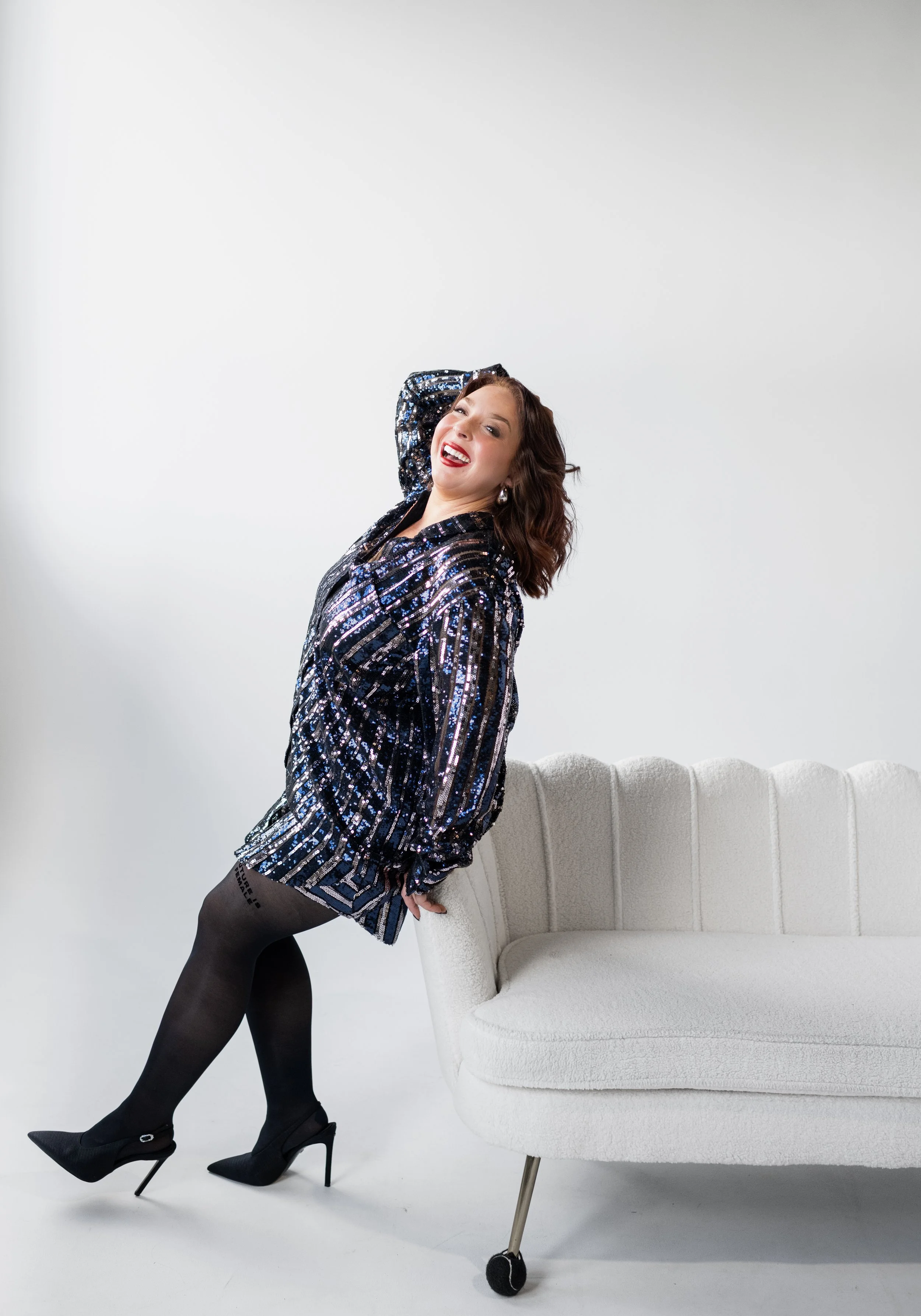 A woman with wavy brunette hair, wearing a shimmery, patterned blazer, black tights, and high heels, leans against a white, modern sofa with one hand behind her head, smiling confidently in a bright studio with a plain white background.