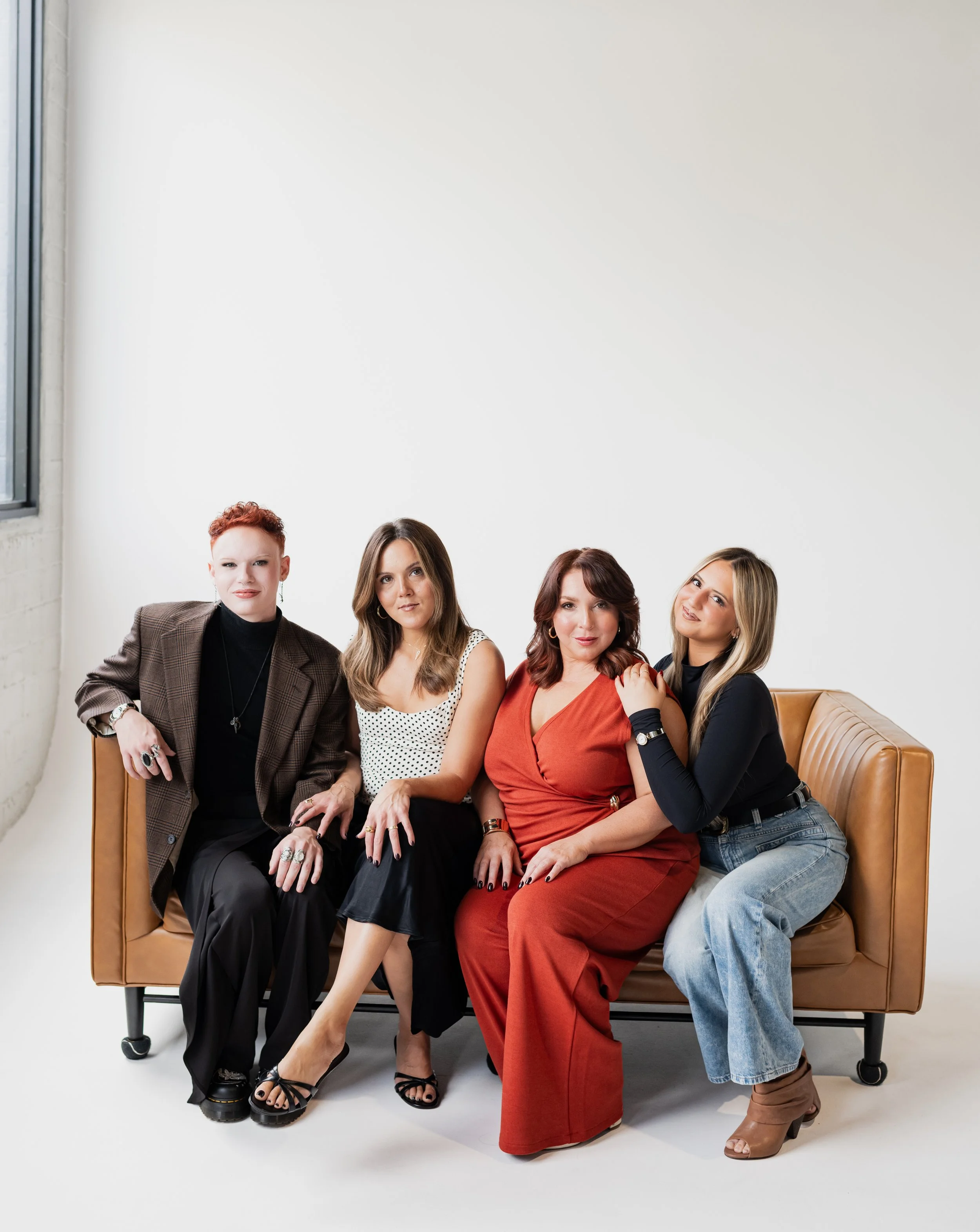 Four women sitting on a tan leather sofa in a studio with a white background, casually dressed with jewelry and fashionable clothing.