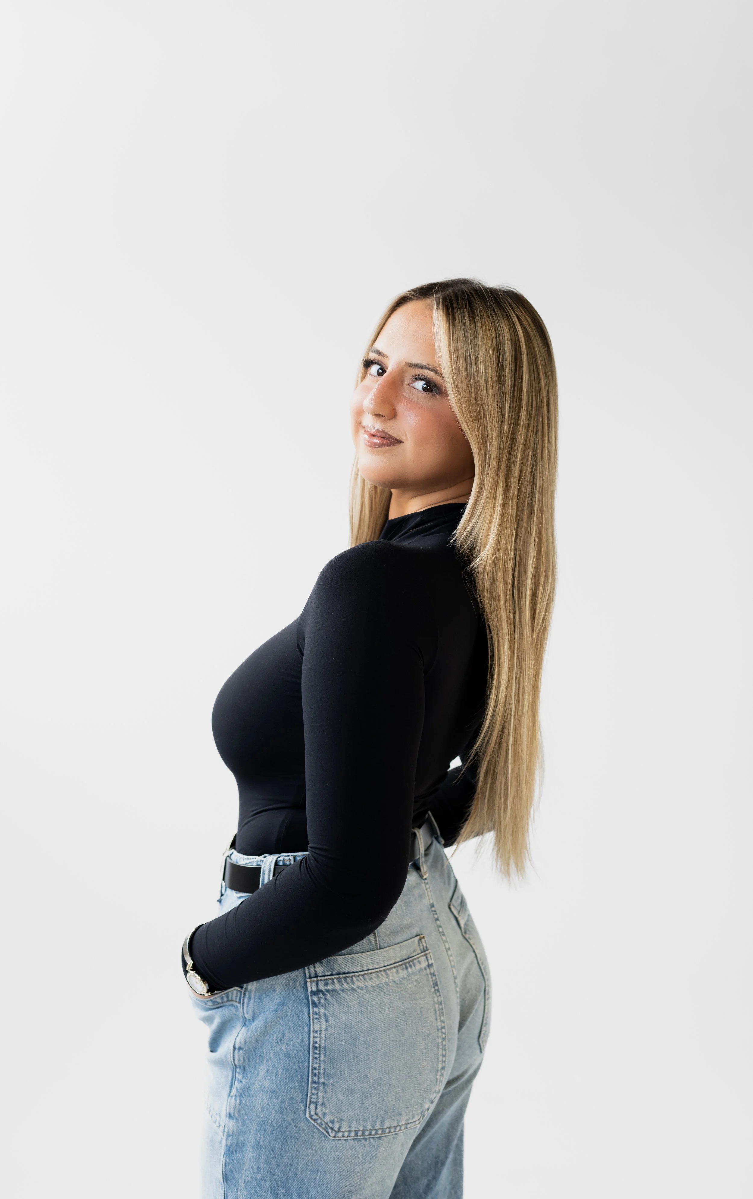 A young woman with long blond hair wearing a black long-sleeve top and light-washed jeans, looking over her shoulder with a slight smile, against a plain white background.
