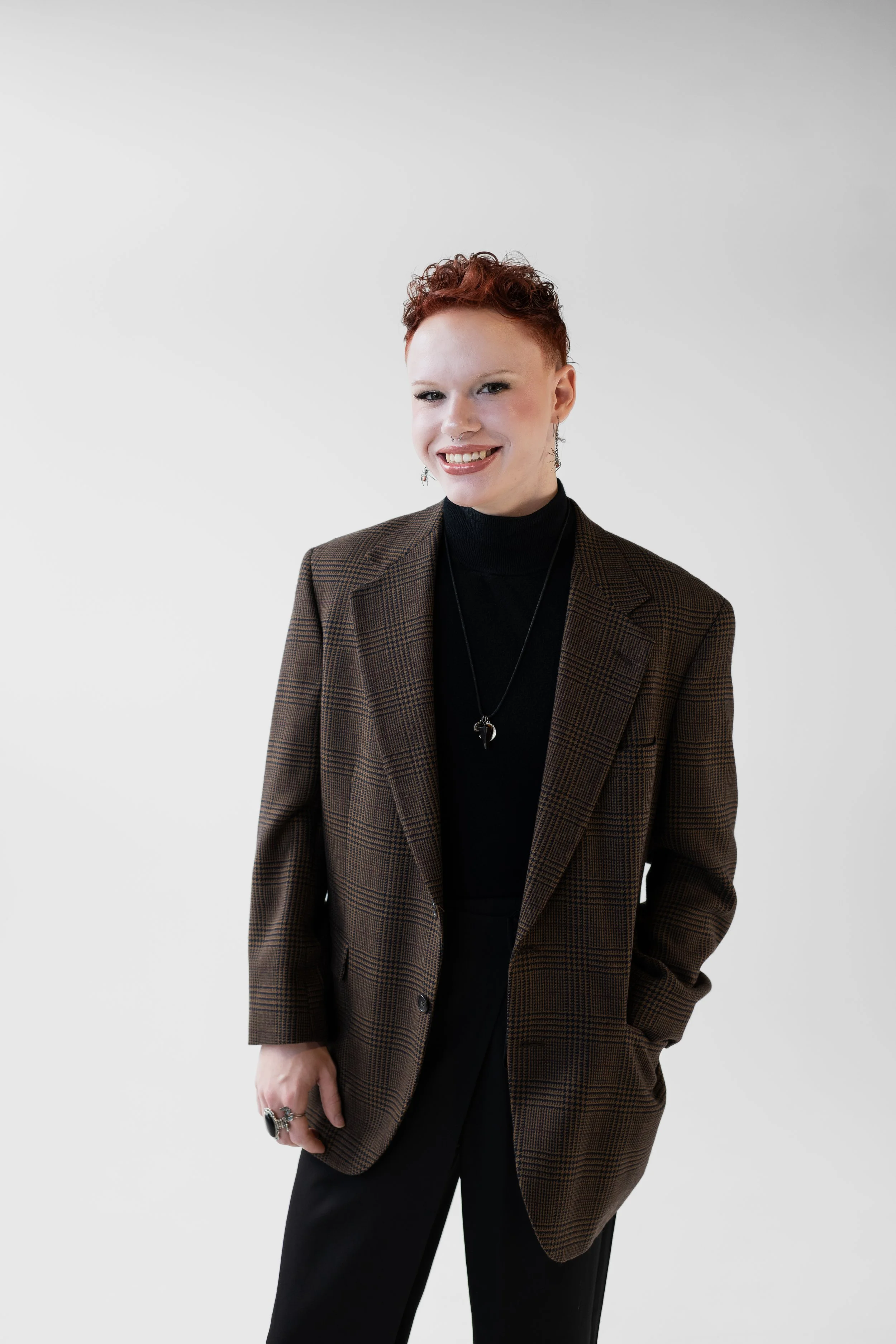 A woman with short curly red hair wearing a black turtleneck, brown plaid blazer, and black pants, smiling against a plain white background.