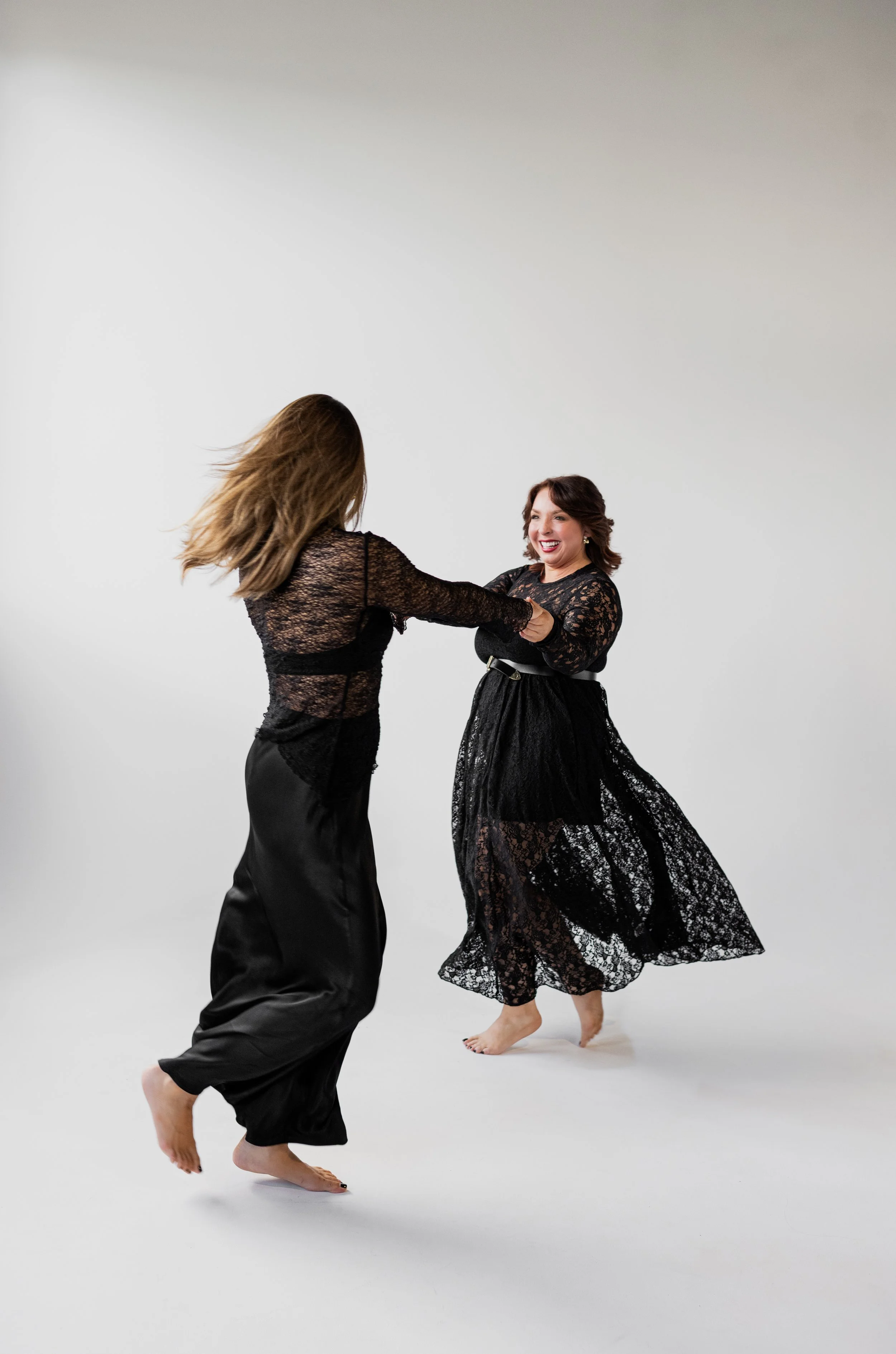 Two women in black lace dresses dance together in a white studio, smiling and holding hands.