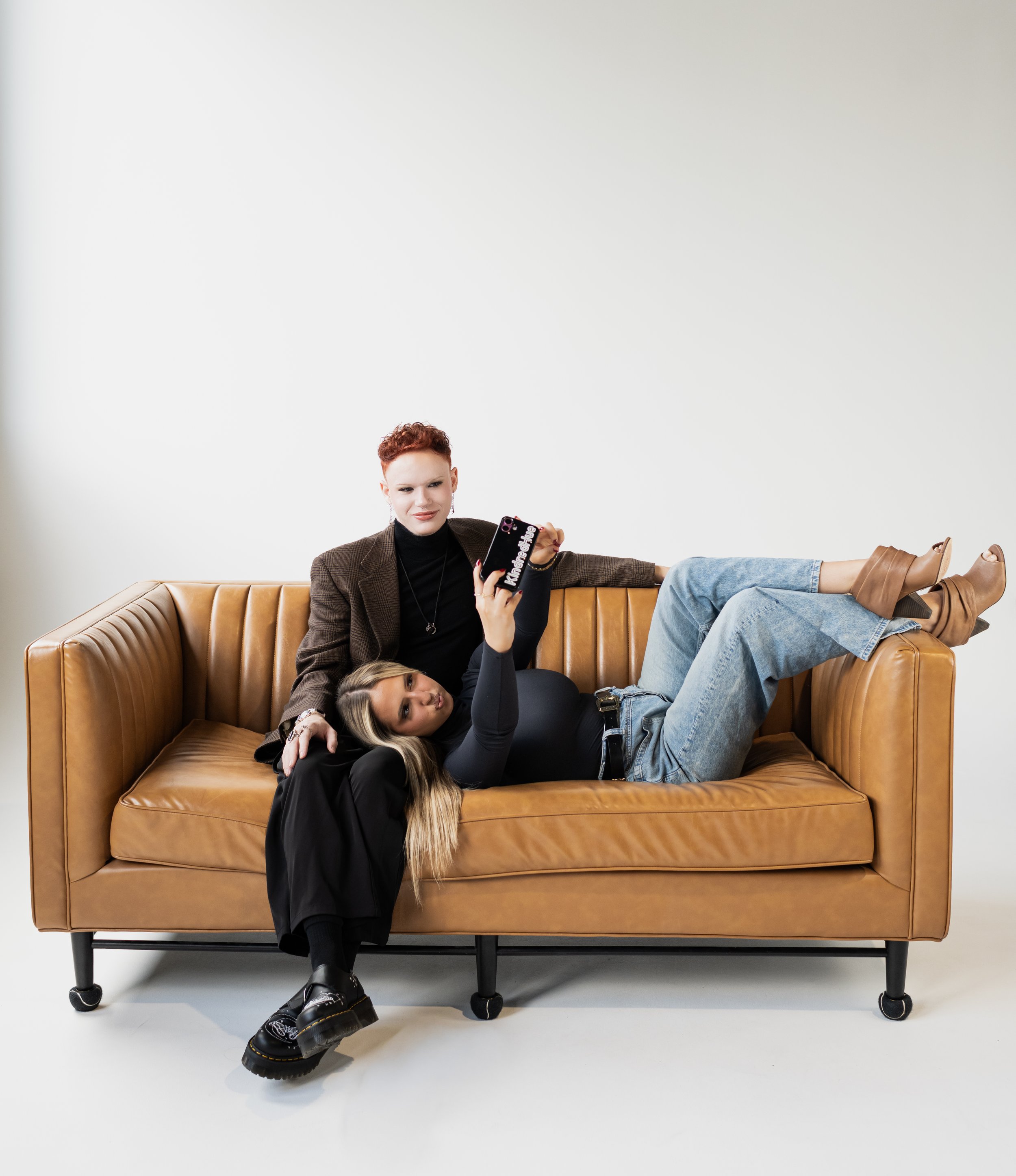 Two women relaxing on a brown leather sofa, one lying down and taking a selfie with a phone, the other sitting behind her with one arm resting on the sofa back.