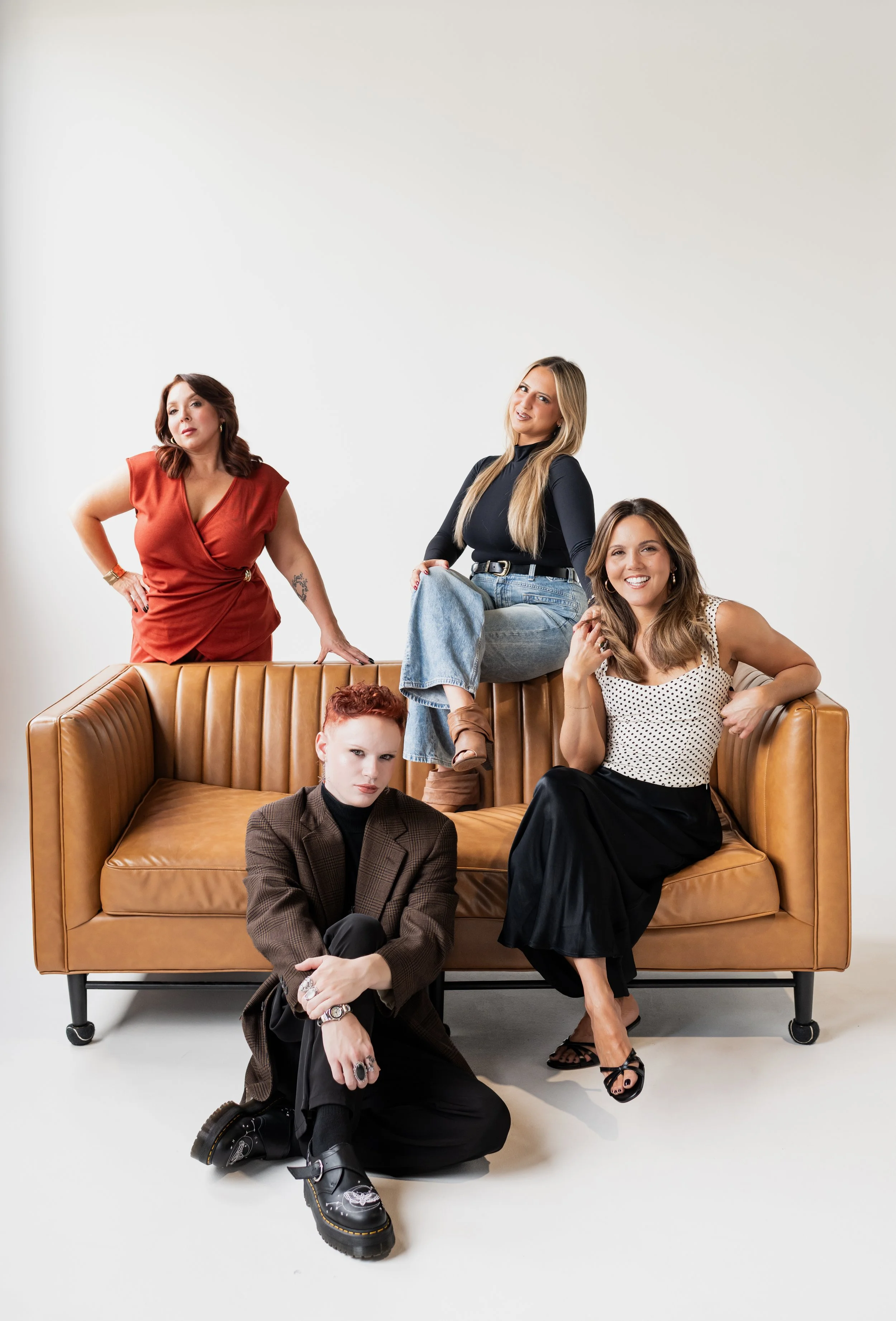 Four women posing around a brown leather sofa against a plain white background.