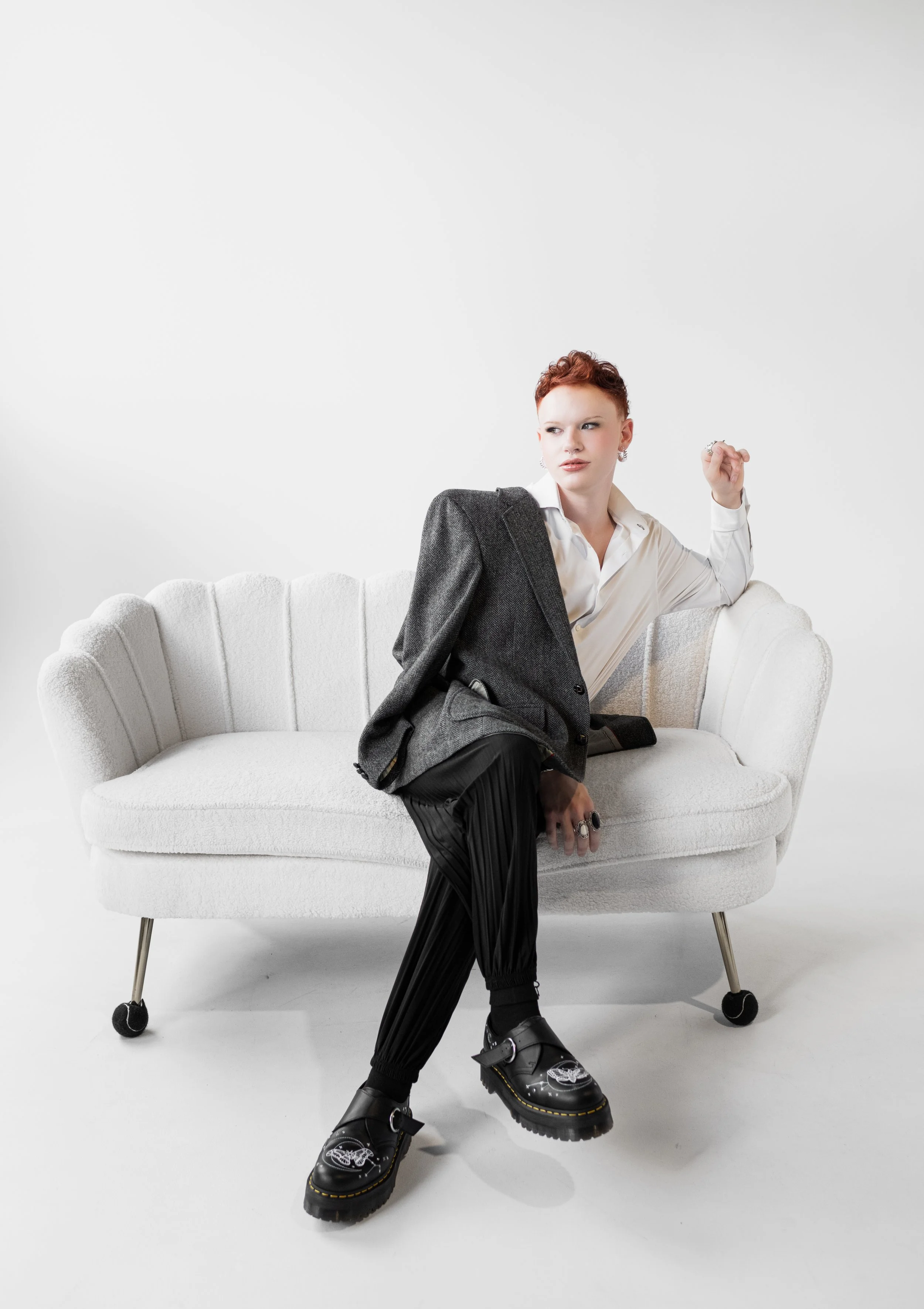 A woman with short red hair sitting on a white vintage style couch, wearing a white shirt, black pleated pants, black chunky shoes, and a gray blazer draped over her shoulder, posed with one arm resting on the back of the couch and looking to the sid