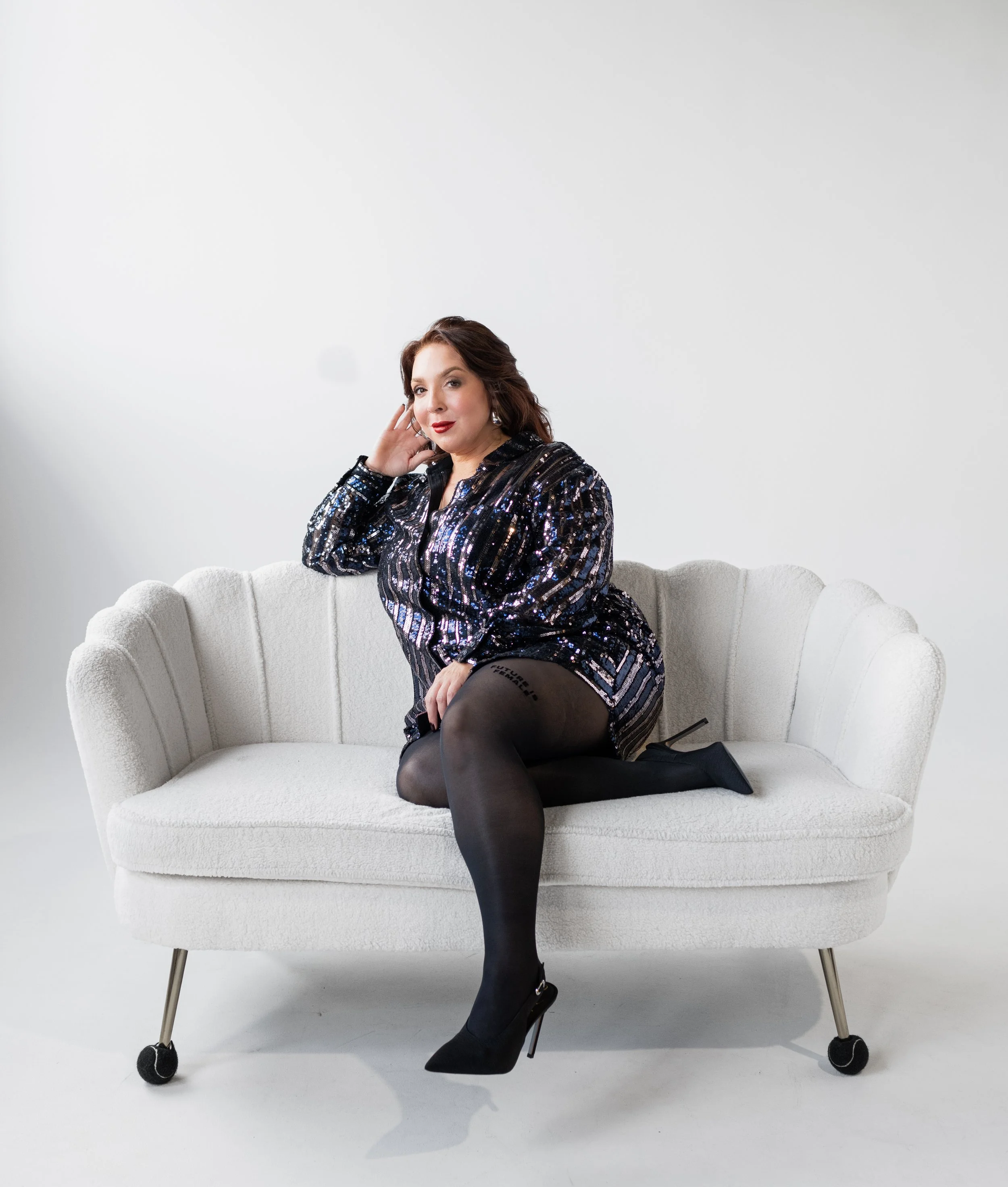A woman sitting on a white vintage style sofa with curved armrests and black legs on small wheels, wearing a black sequined dress and black high heels, posing with her right hand touching her face, against a plain white background.