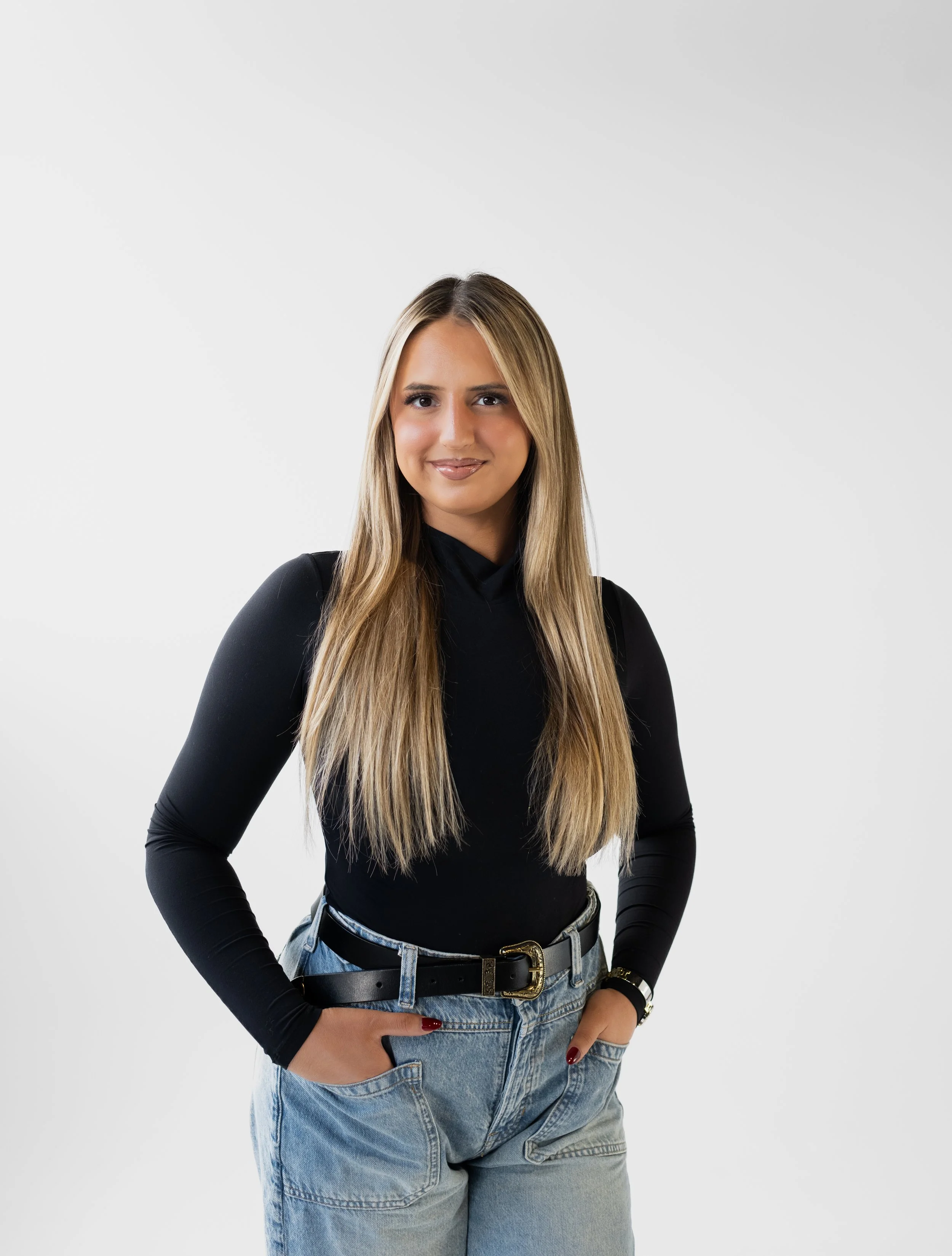A young woman with long blonde hair, wearing a black long-sleeve top, light blue jeans, and a black belt, standing against a plain white background.