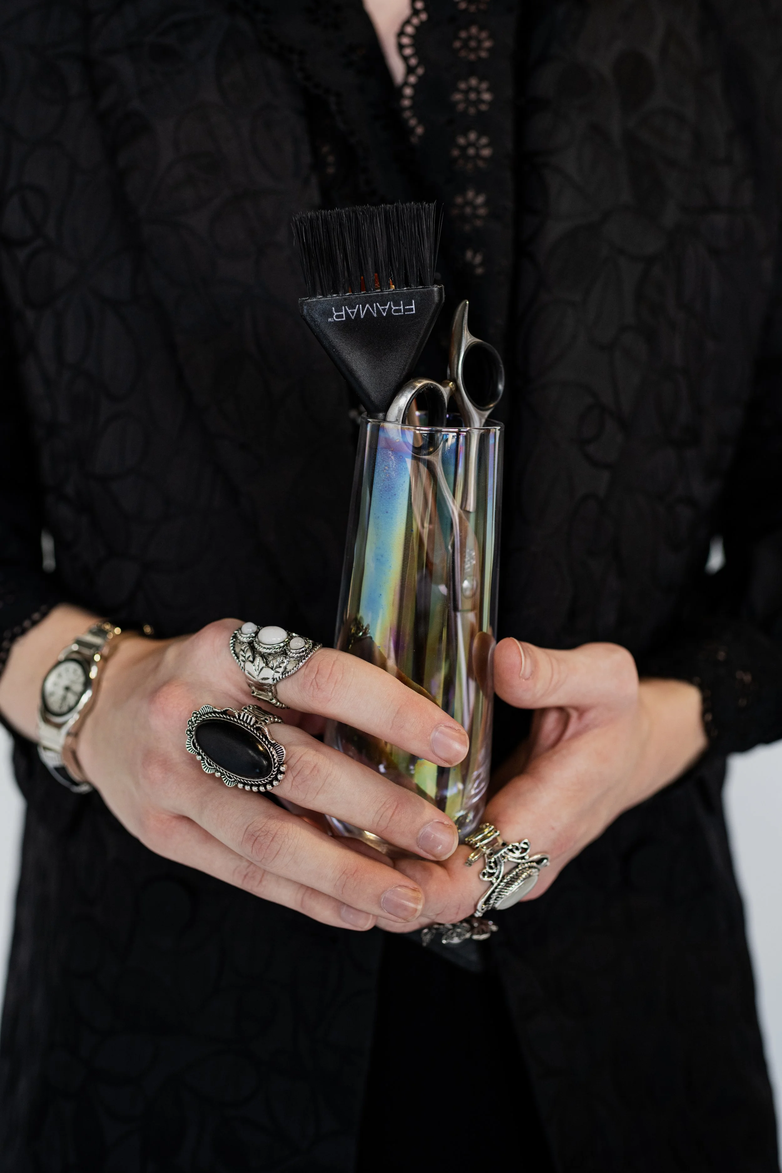 Person holding a glass vase containing hair cutting tools, including scissors, a comb, and a small brush, with a black quilted jacket and rings on their fingers.