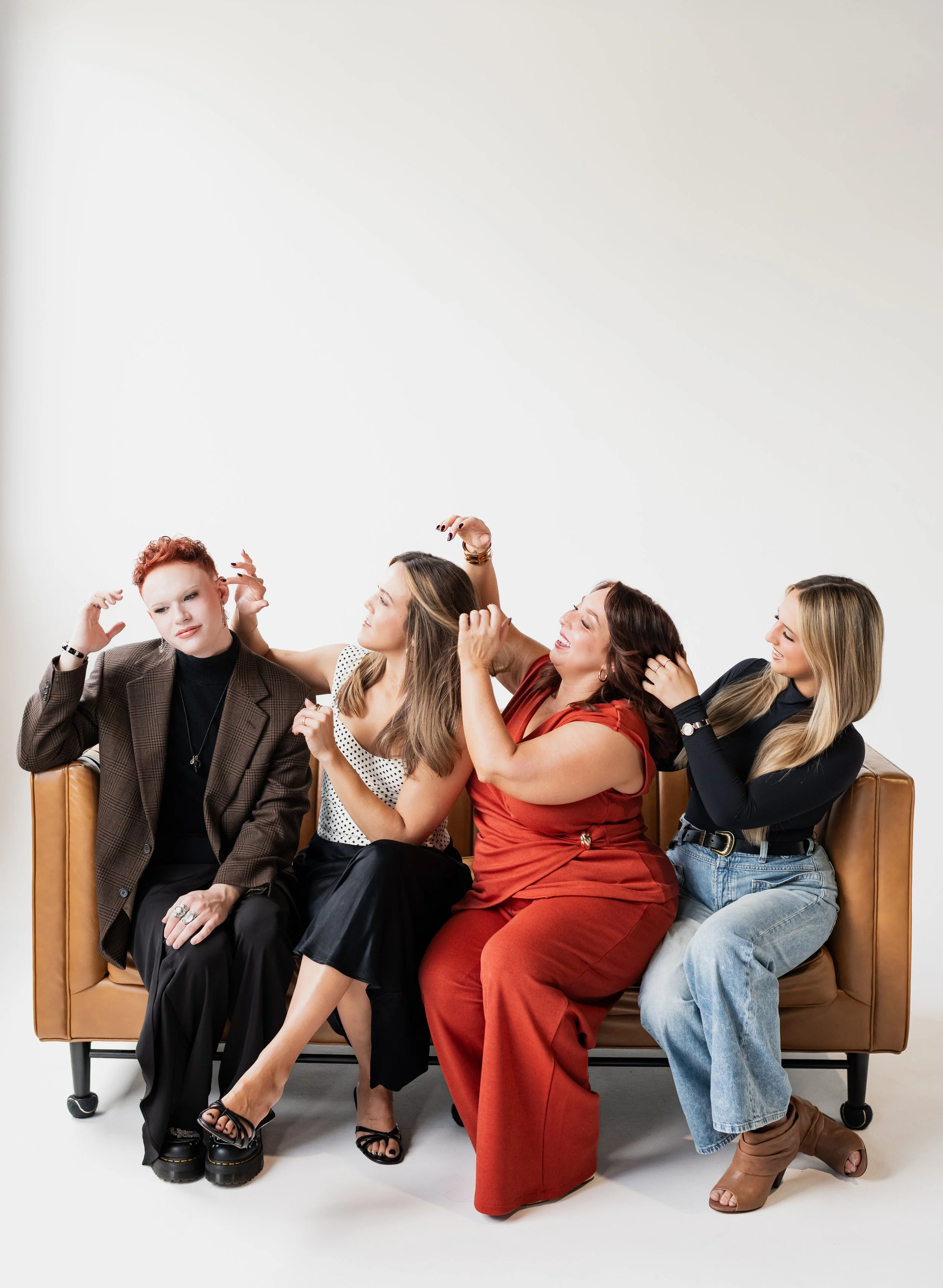Four women sitting on a tan sofa, making playful facial expressions and gestures, against a plain white background.