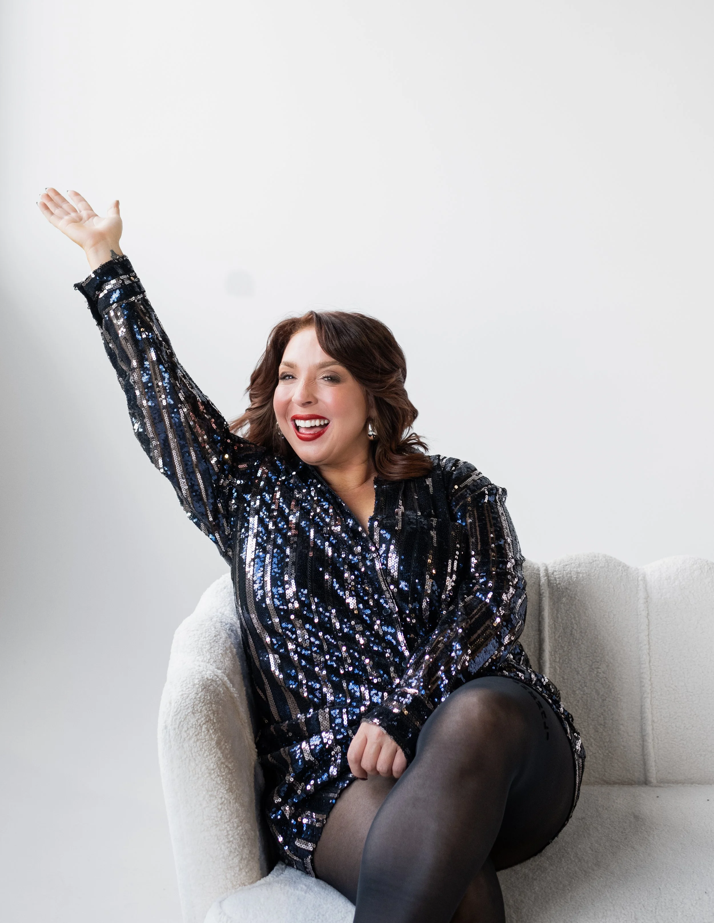 A woman with brown hair and red lipstick wearing a black sequined dress, sitting on a white couch, smiling, and raising her right arm.