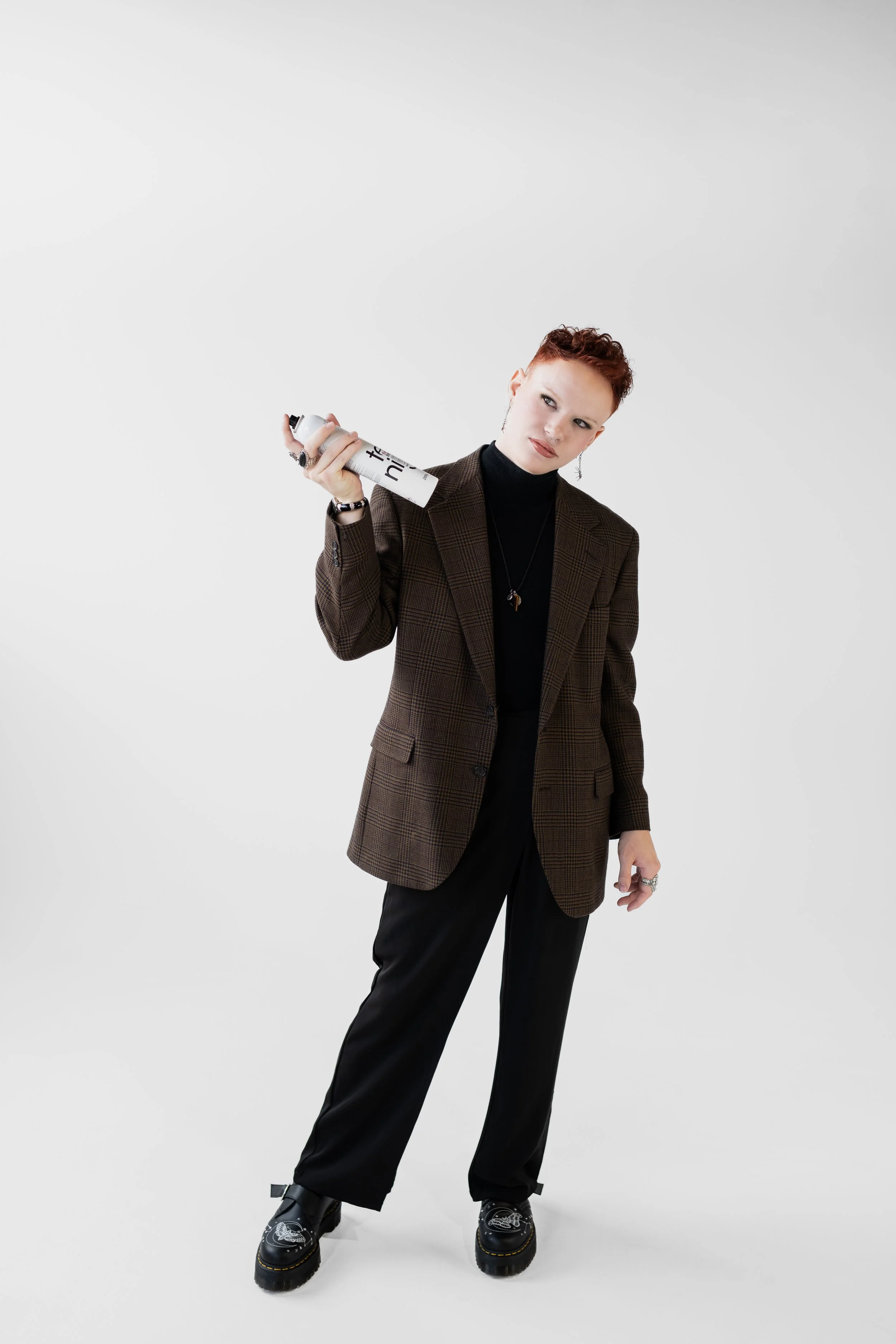 A woman with short red hair dressed in a plaid blazer, black turtleneck, and black pants, holding a spray paint can over her shoulder, standing on a plain white background.