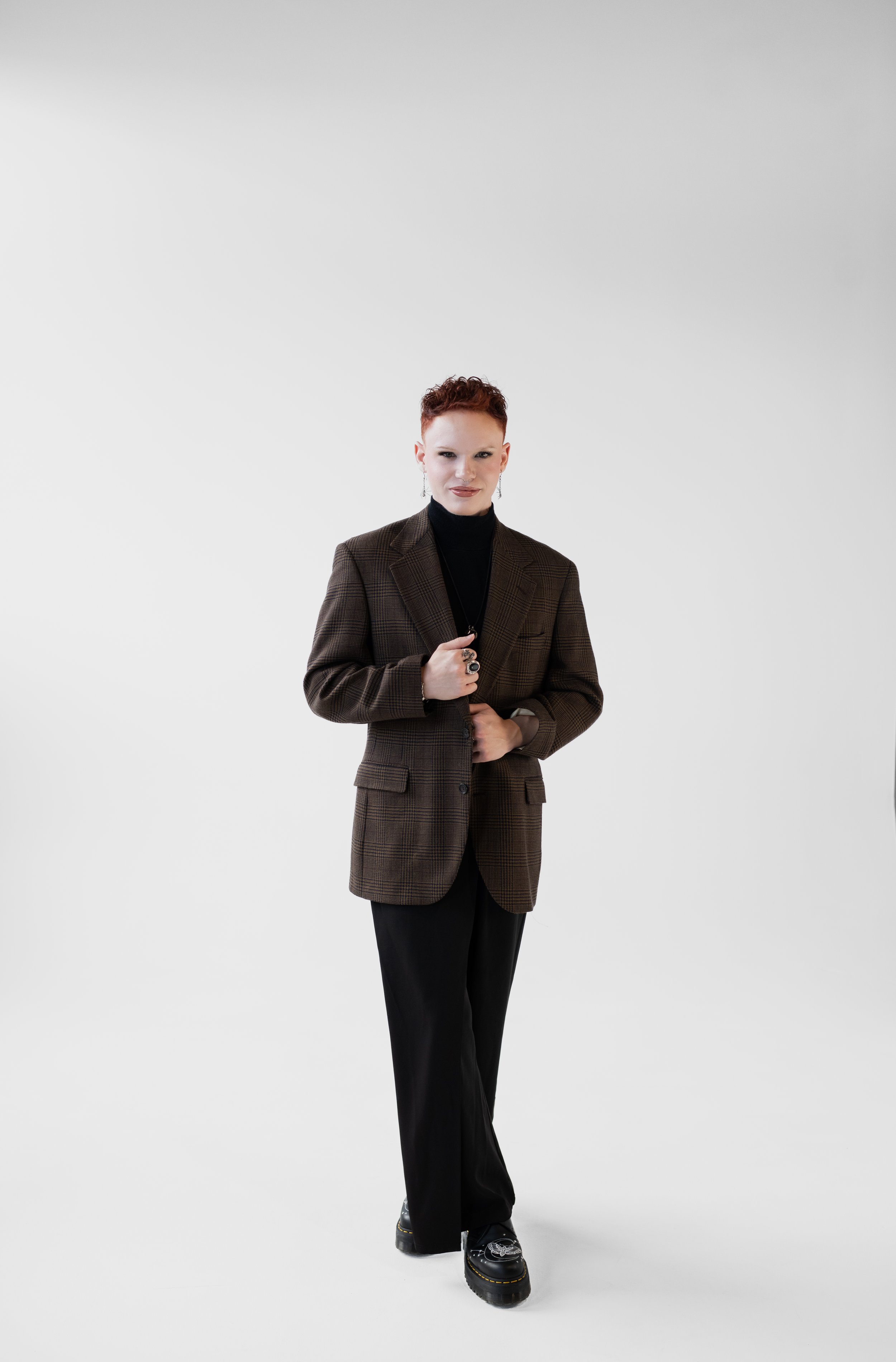 A woman with short reddish hair wearing a brown plaid blazer, black turtleneck, black wide-leg pants, and black platform shoes with a pattern, standing against a plain white background.