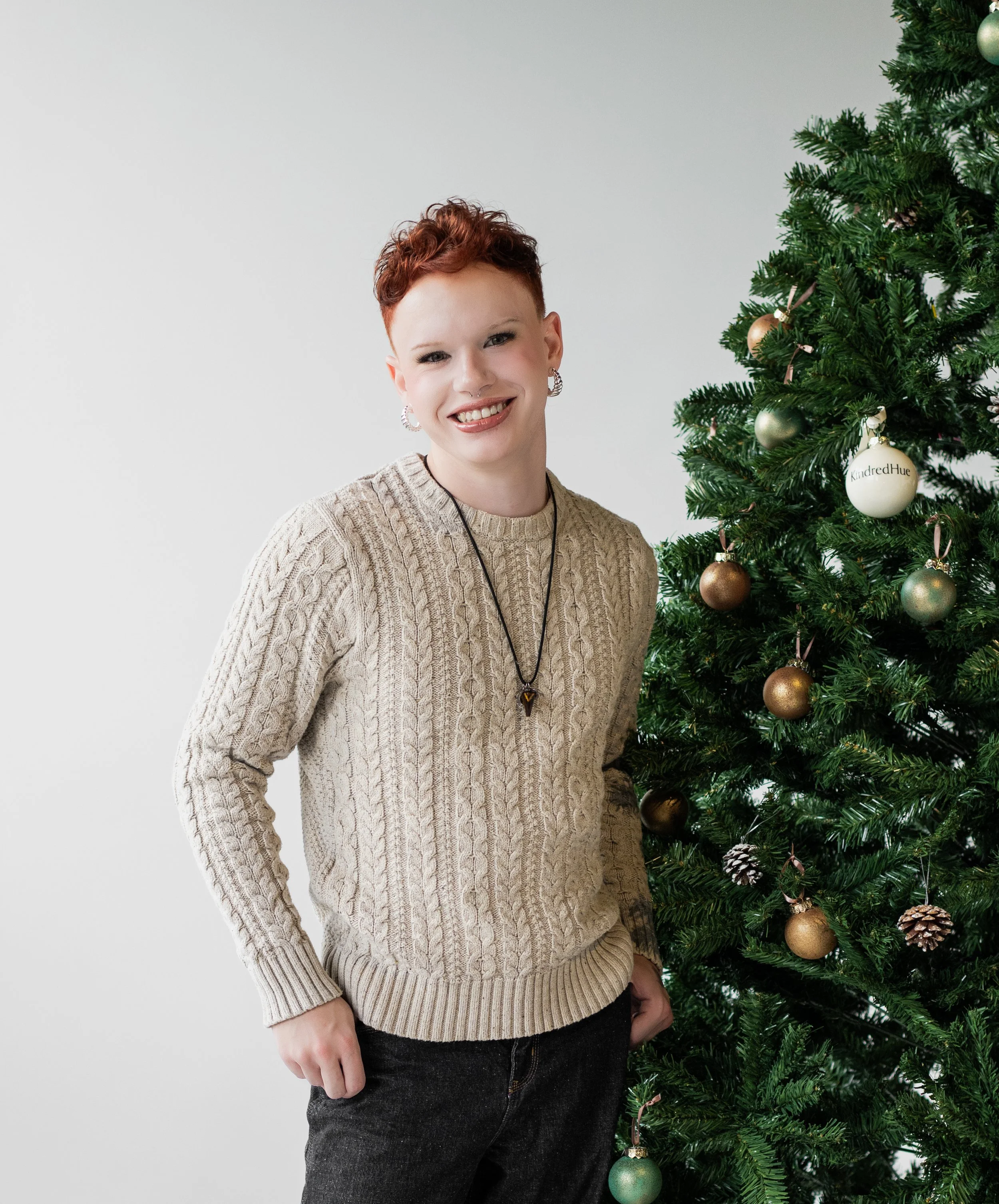 A smiling person with short red hair standing next to a decorated Christmas tree, wearing a beige cable-knit sweater, black pants, and jewelry.