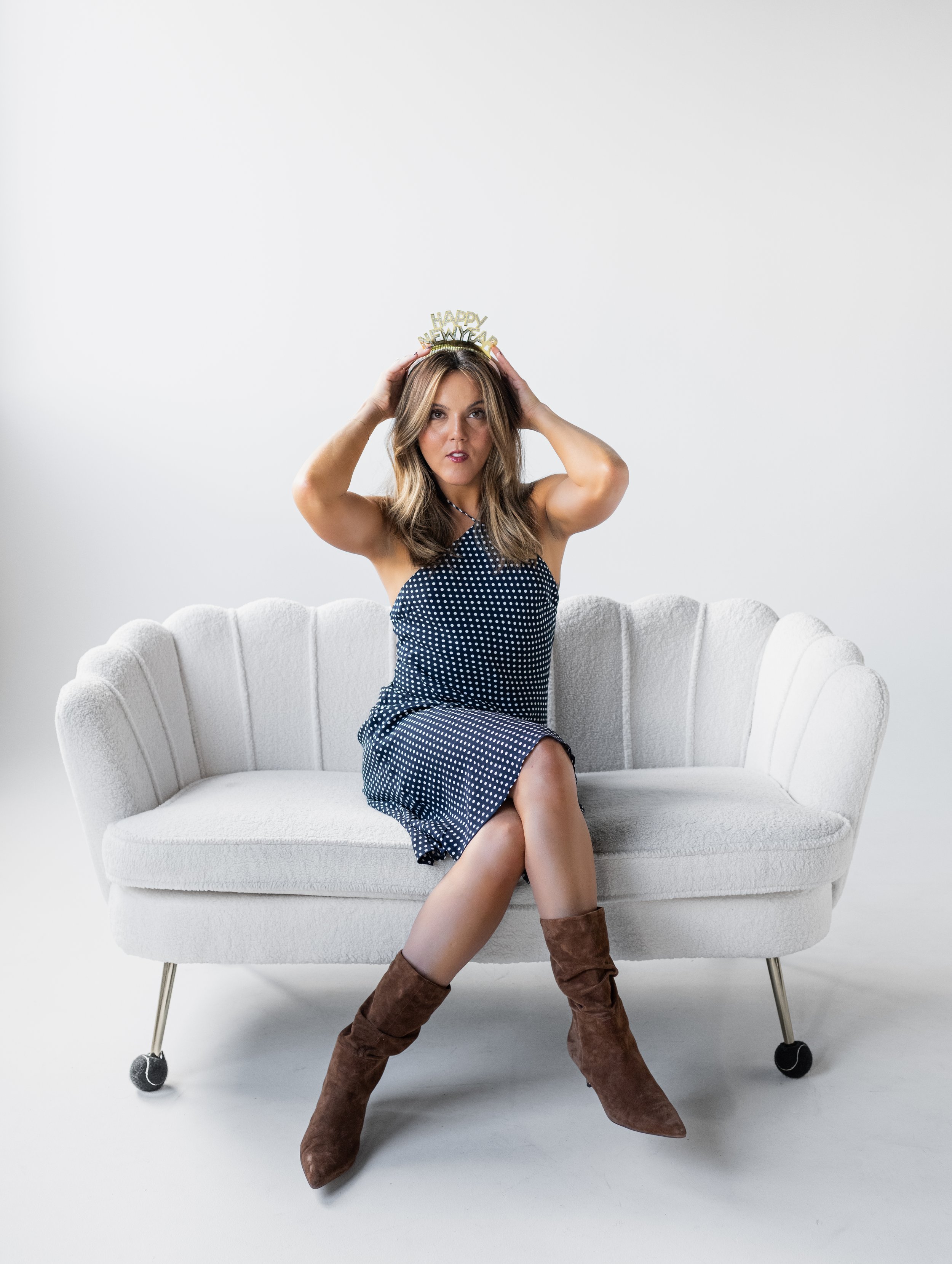 A woman sitting on a white sofa, wearing a polka dot dress and brown boots, holding a 'Happy New Year' headband behind her head.