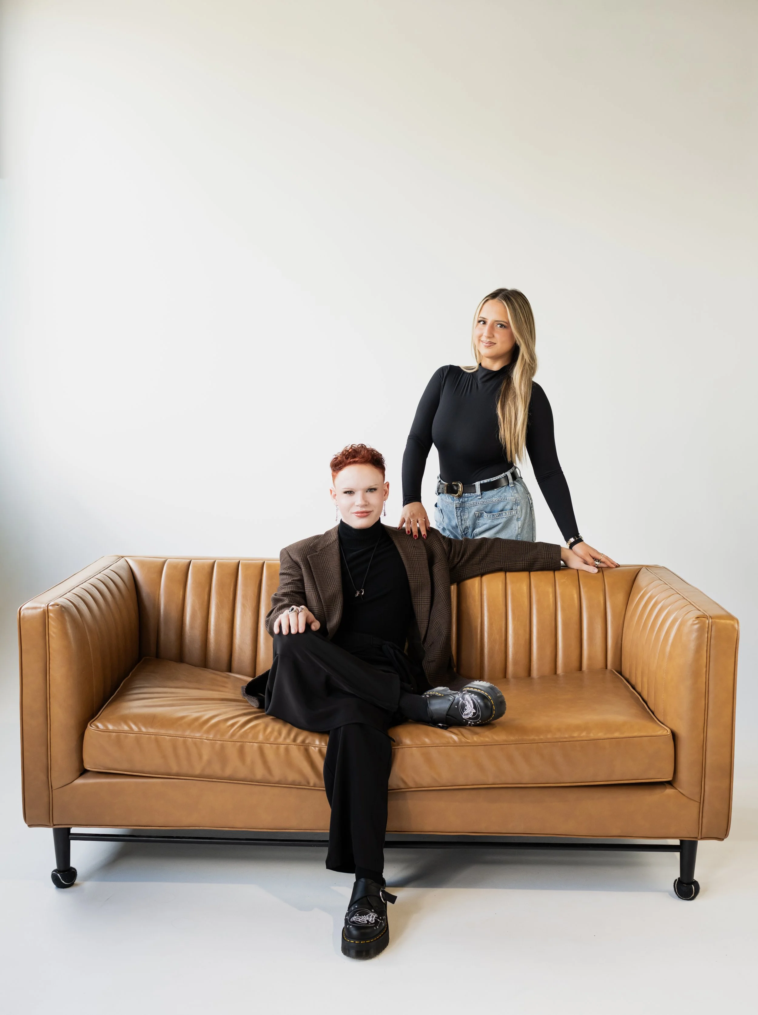 Two women, one sitting on a tan leather couch and the other standing behind it, in a studio with a plain white background.