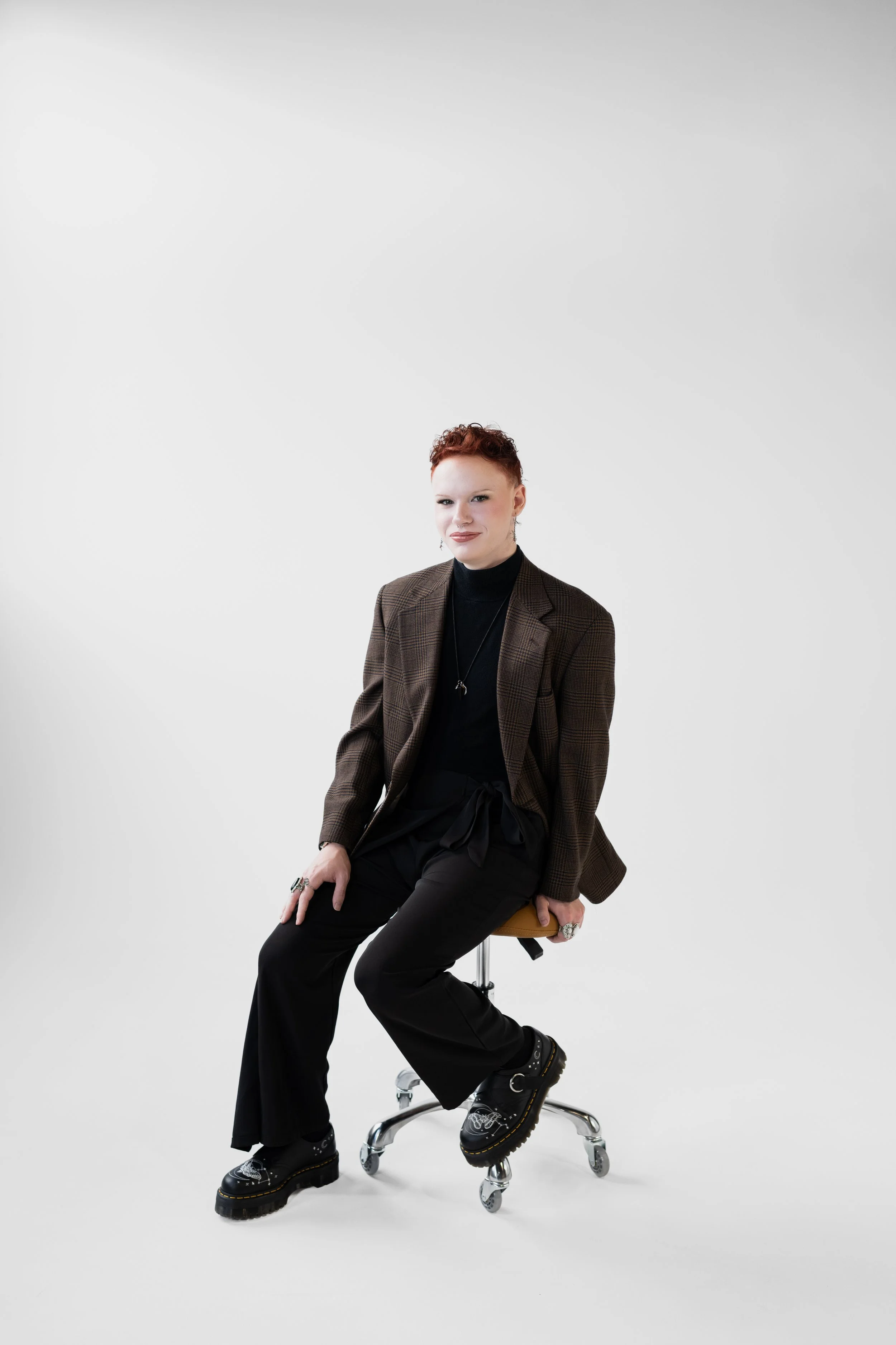 A woman with red hair sitting on a stool in a studio with a white background, wearing a brown plaid blazer, black turtleneck, black pants, and black platform shoes, accessorized with rings and necklaces.