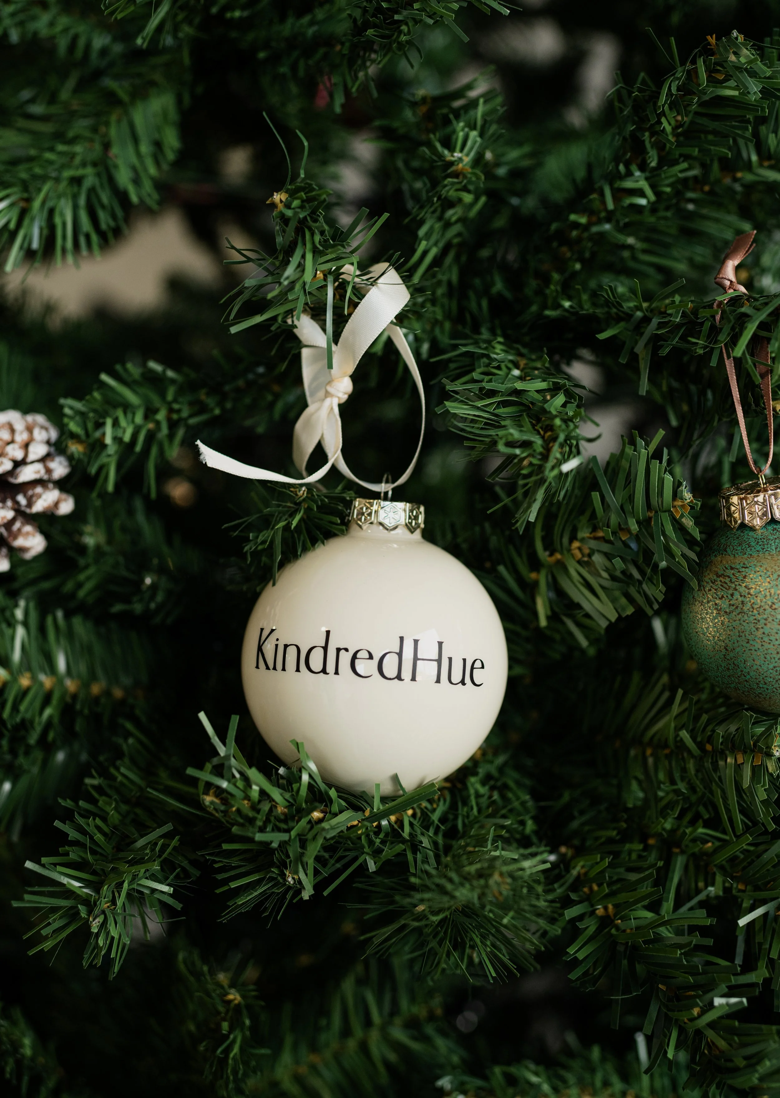 A white Christmas ornament hanging on a Christmas tree with the text "KindredHue" written on it.