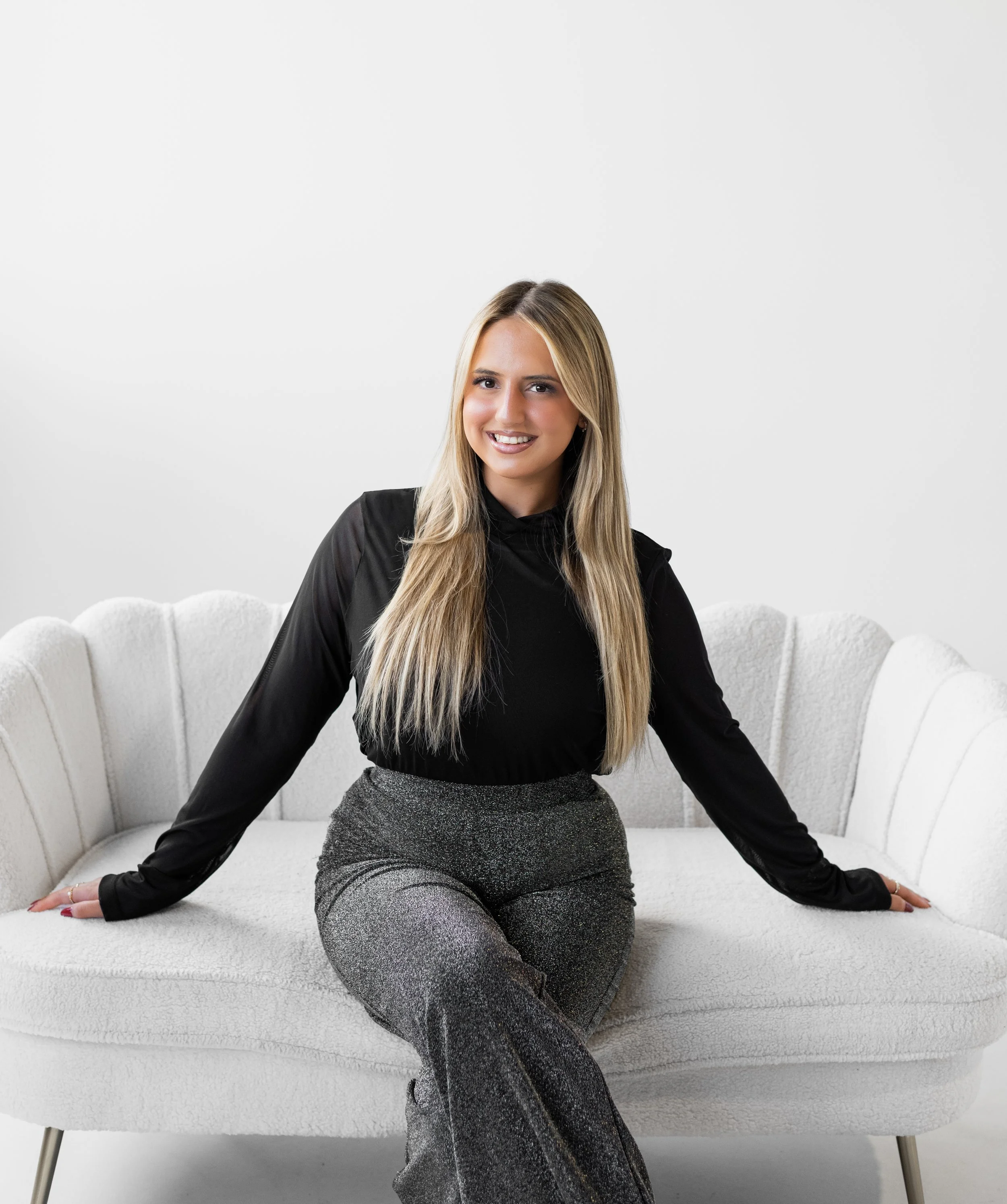 A young woman with long blonde hair, wearing a black long-sleeve top and sparkly gray pants, sitting on a white upholstered sofa in front of a plain white background, smiling at the camera.