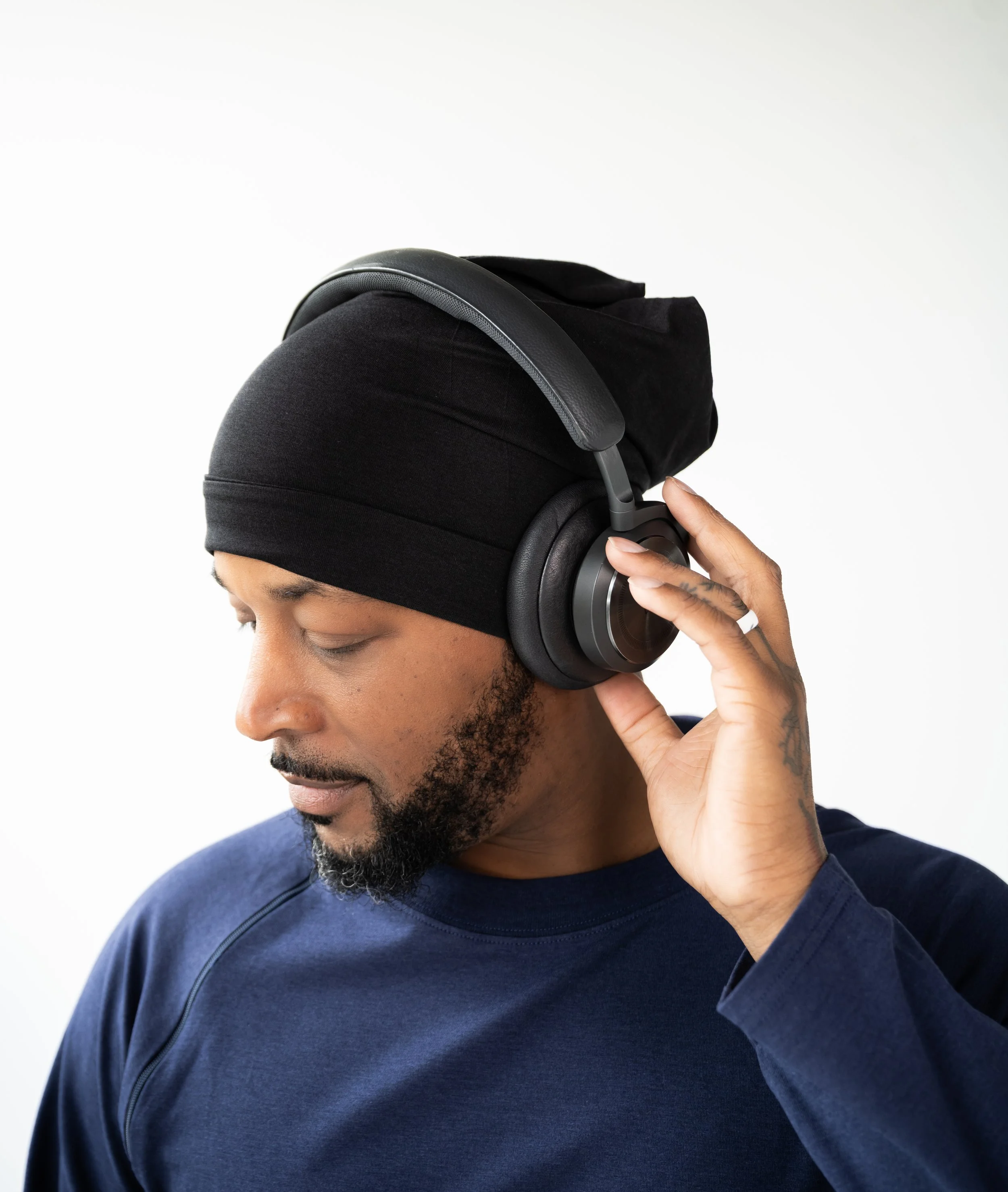A man in a black beanie wearing headphones and a navy blue shirt, with his eyes closed and one hand adjusting the headphones.