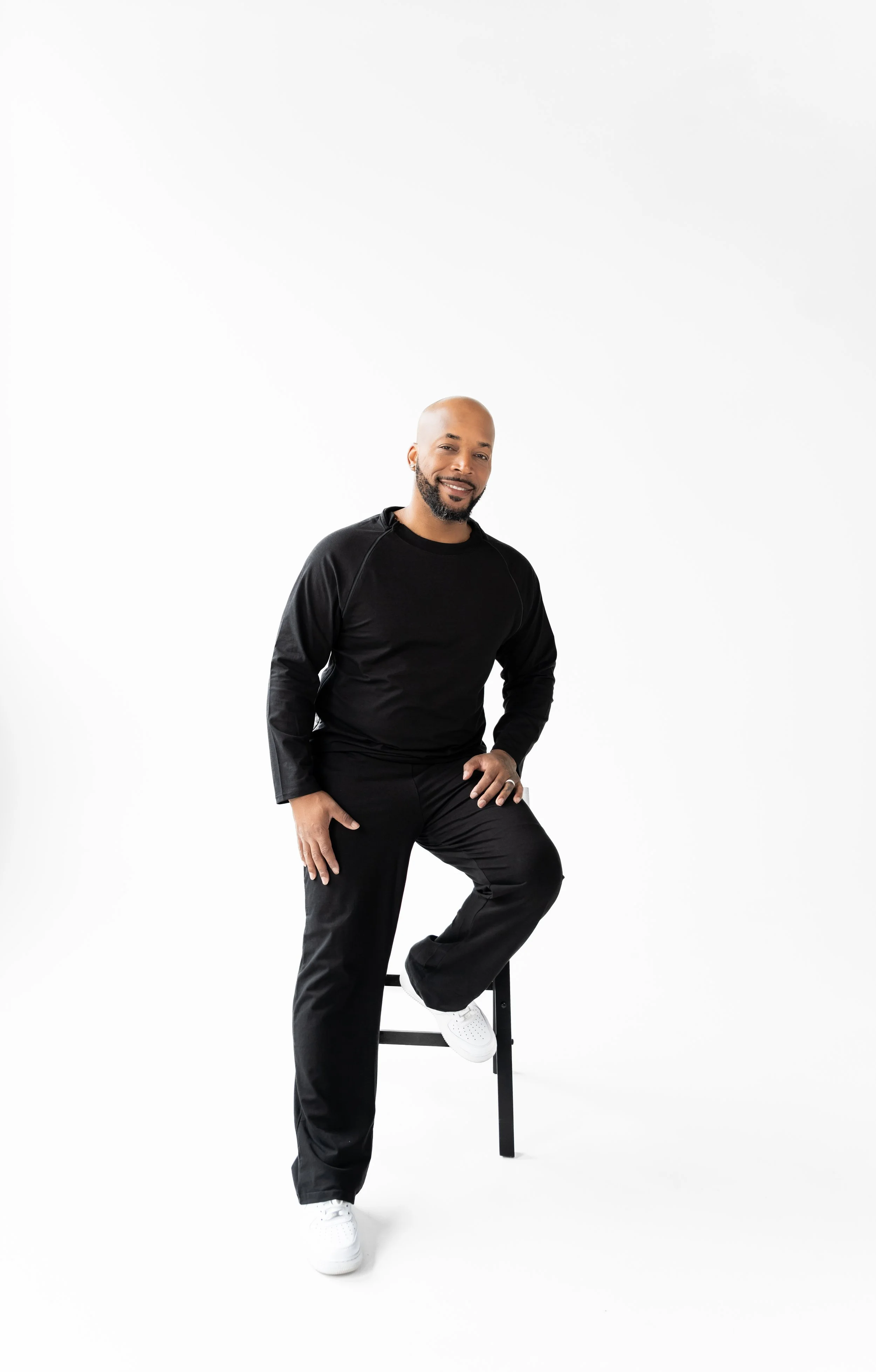 A man with a beard and bald head, wearing a black long-sleeve shirt and black pants, smiling and resting one foot on a black stool in a white studio setting.