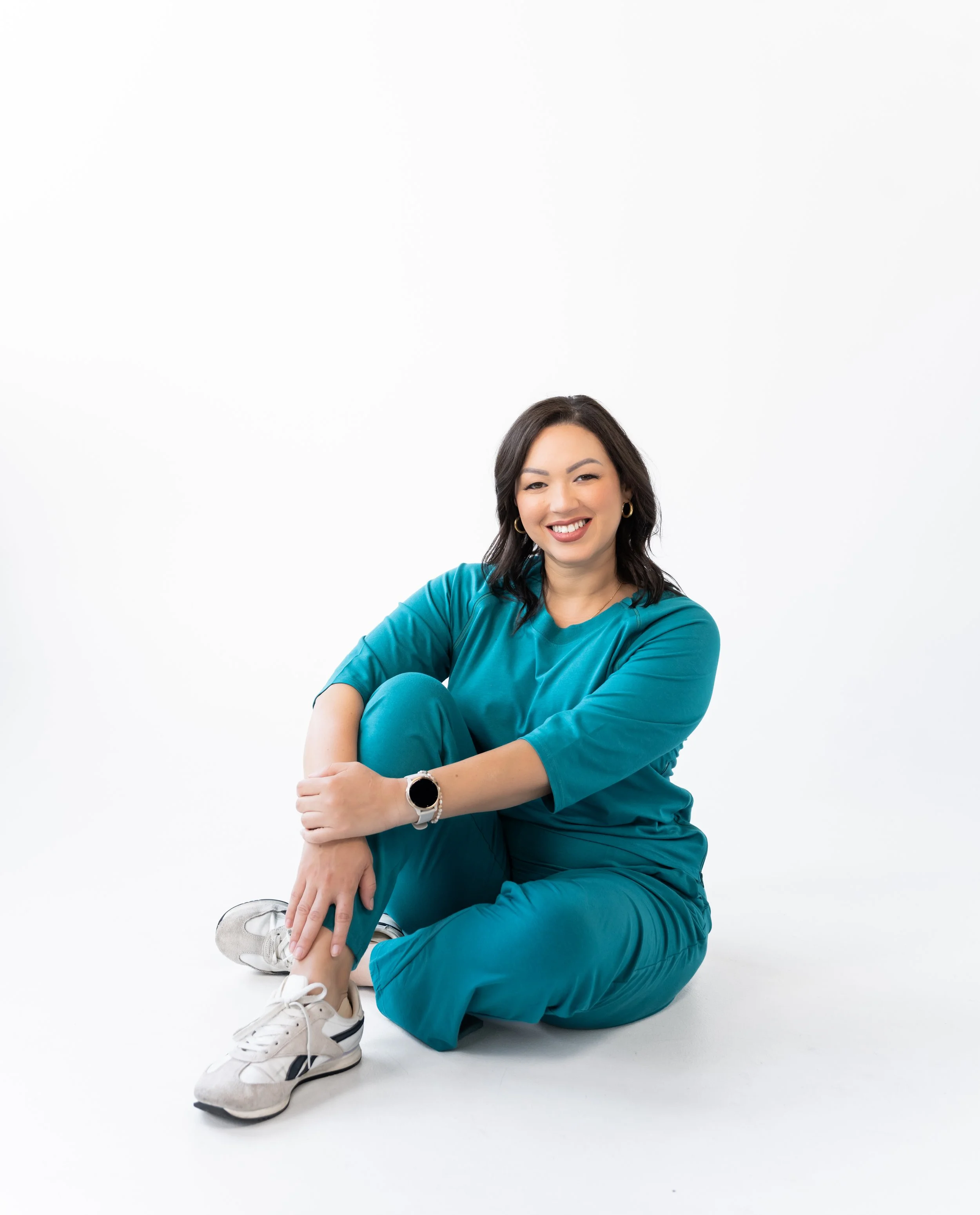 Woman sitting on the floor with crossed legs, smiling, wearing teal scrubs, sneakers, and a smartwatch, against a plain white background.