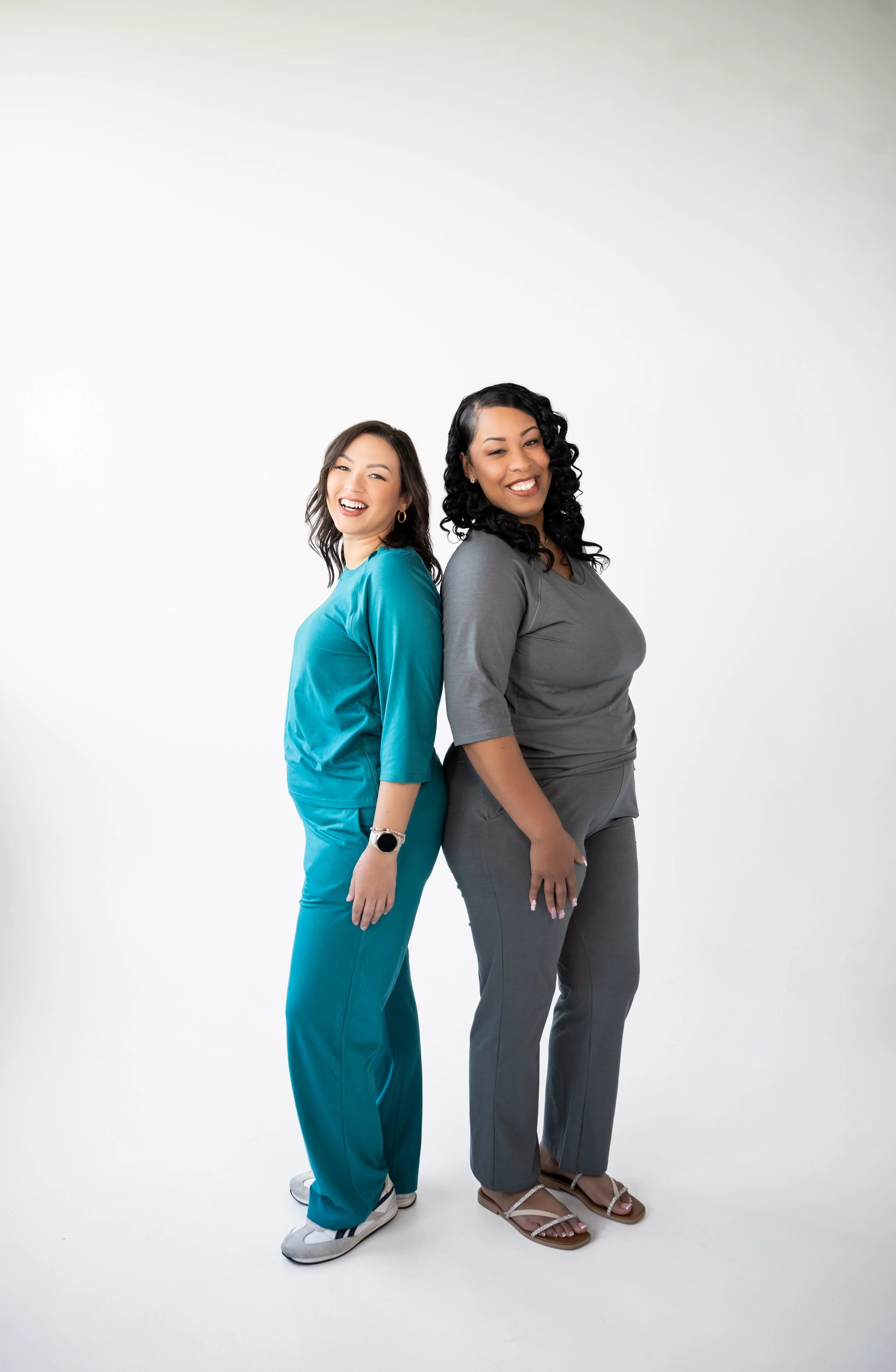 Two women standing back-to-back, smiling, one wearing teal scrubs and the other in gray scrubs, against a white background.