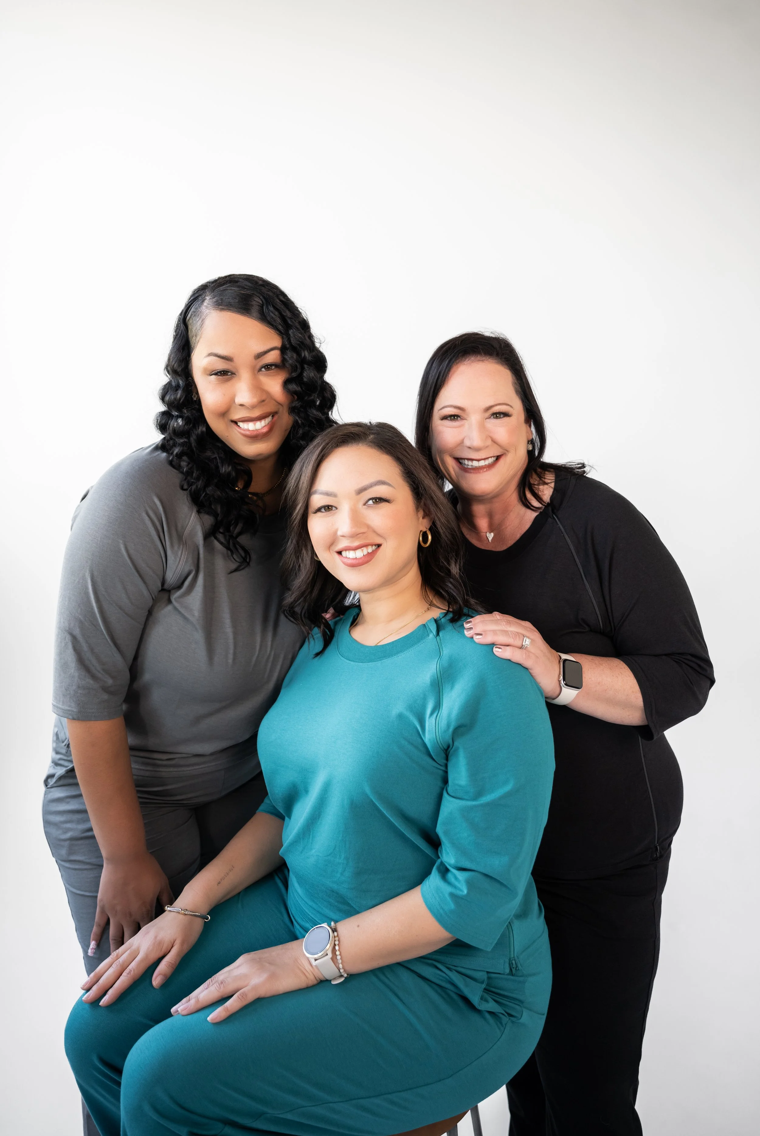 Three women smiling together, with one seated in teal scrubs and two standing behind her, in a studio with a white background.
