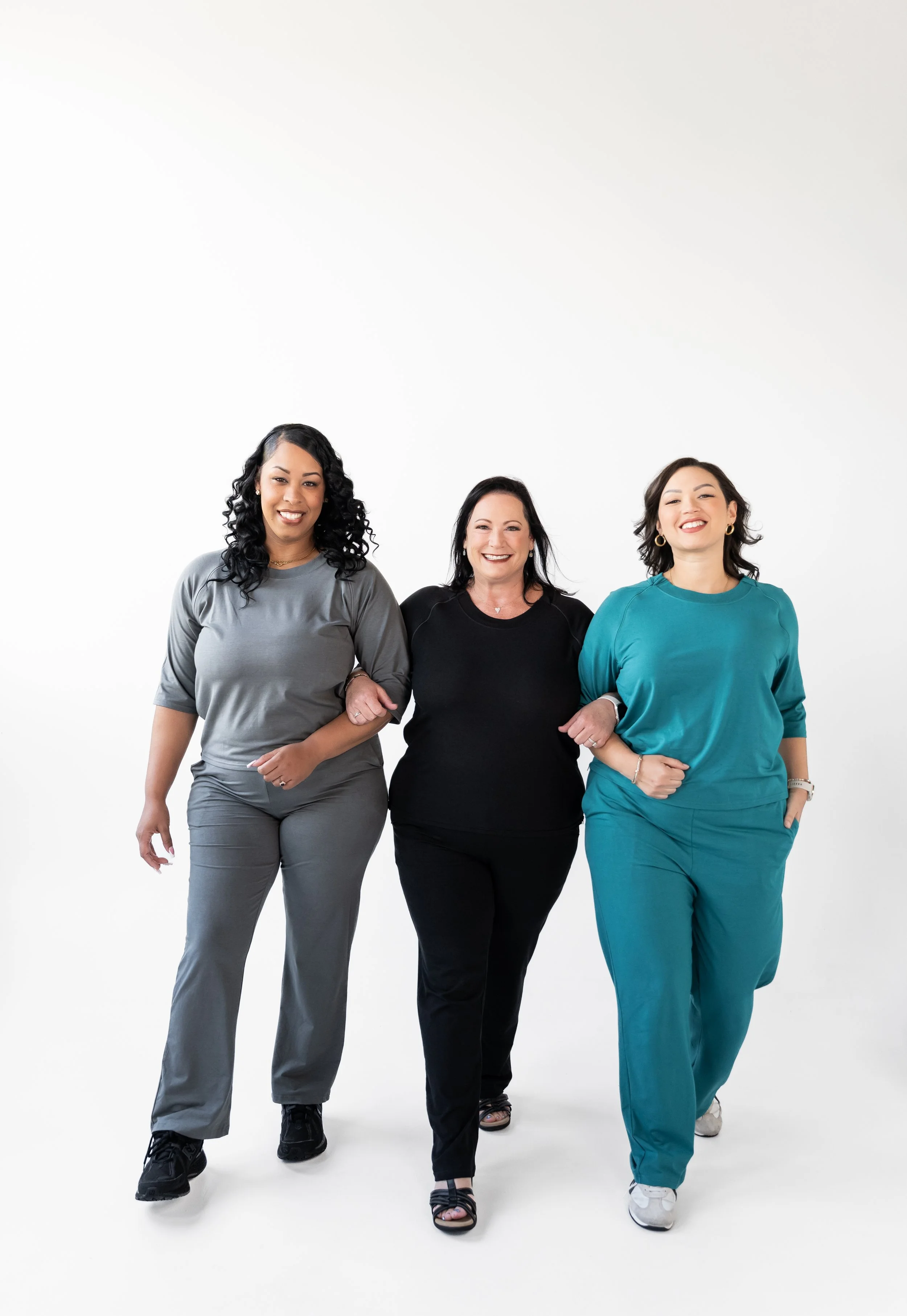 Three women walking together, arm in arm, smiling against a plain white background.