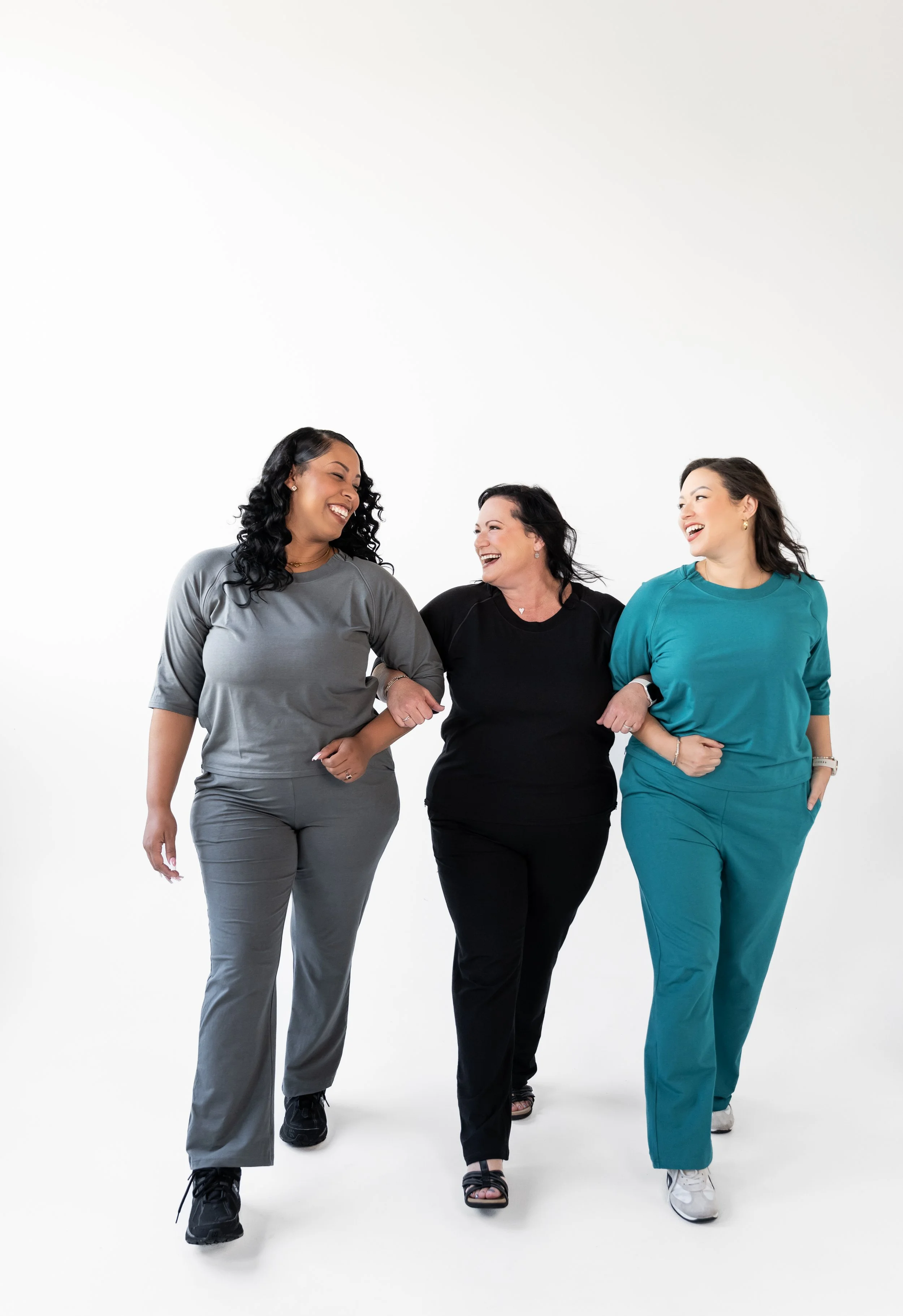 Three women walking arm-in-arm, smiling and laughing, against a plain white background.