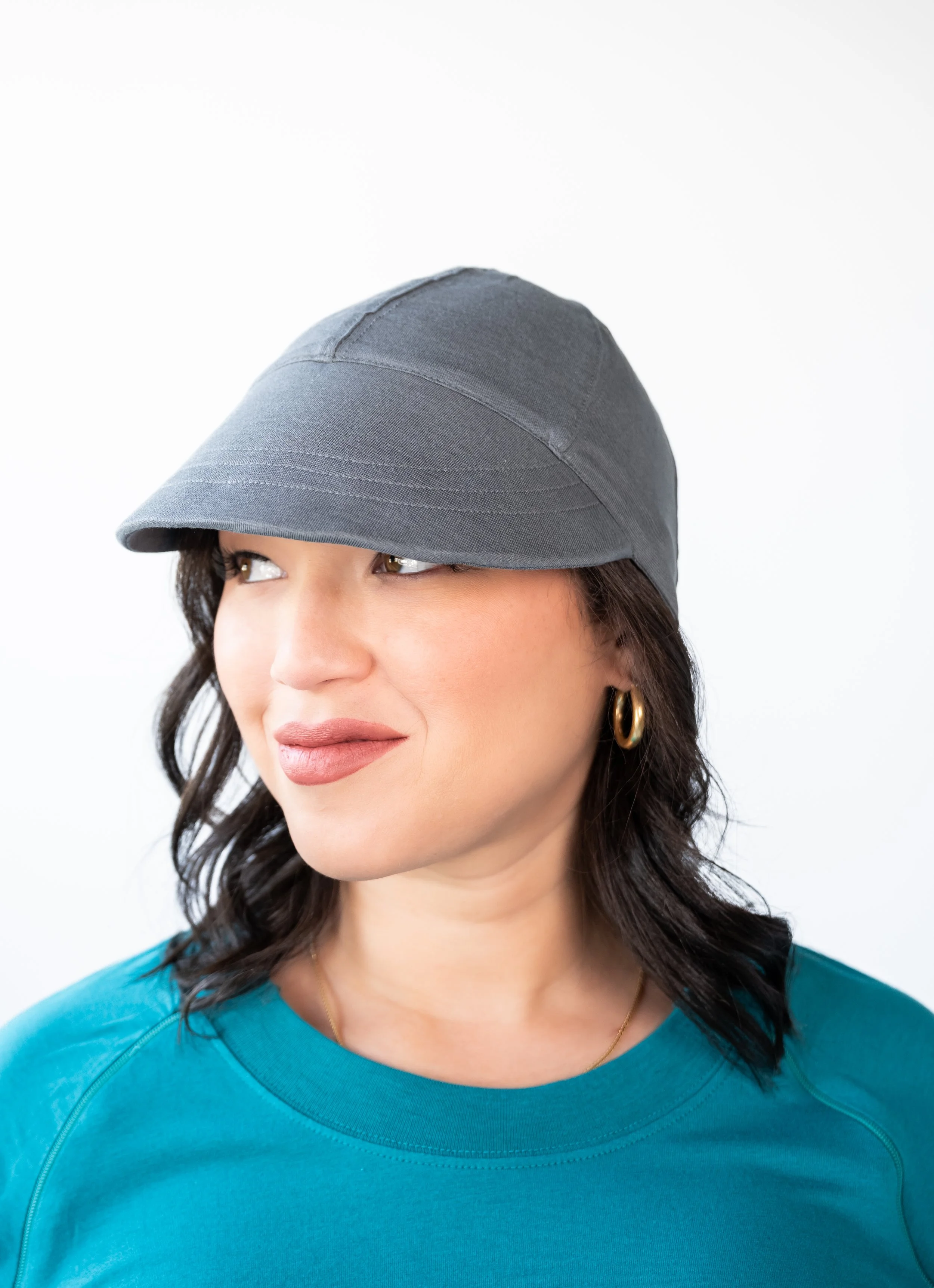 A woman with shoulder-length dark hair wearing gold hoop earrings, a blue top, and a gray cap that covers her eyes, looking slightly to the side against a white background.