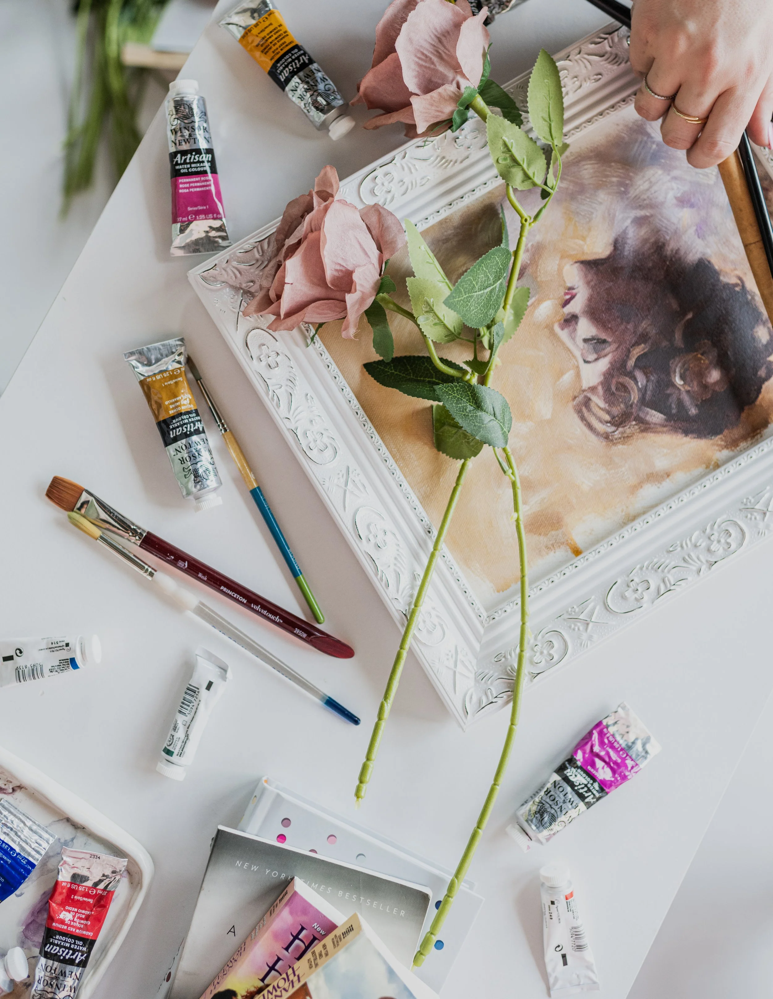 A white table with art supplies, including paint tubes, brushes, and a partially completed floral painting in a decorative white frame. There are two artificial roses on the table and an open book nearby.