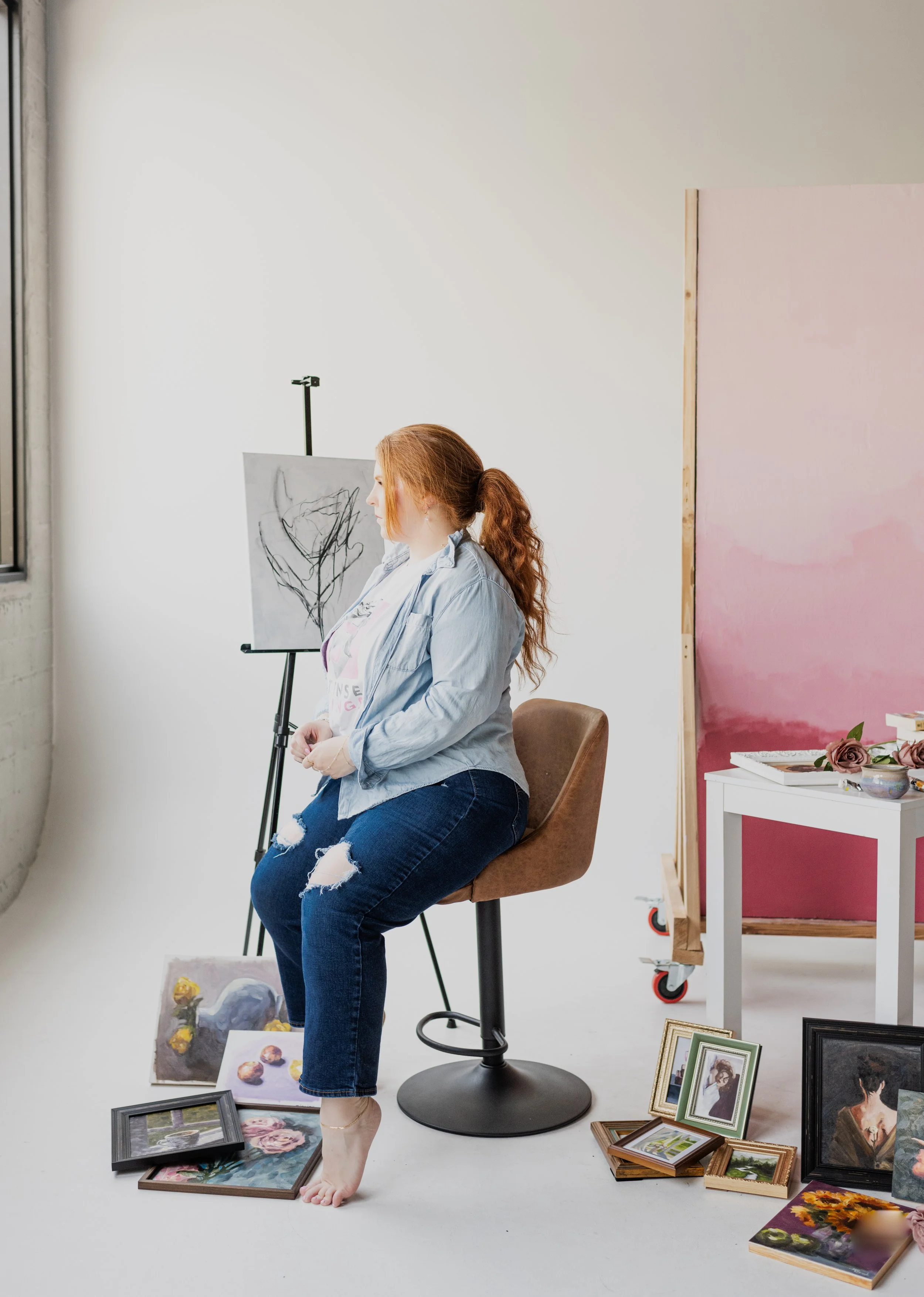 A red-haired woman sitting on a brown stool, surrounded by paintings, framed photos, and art supplies in an art studio with a pink backdrop and a white table.